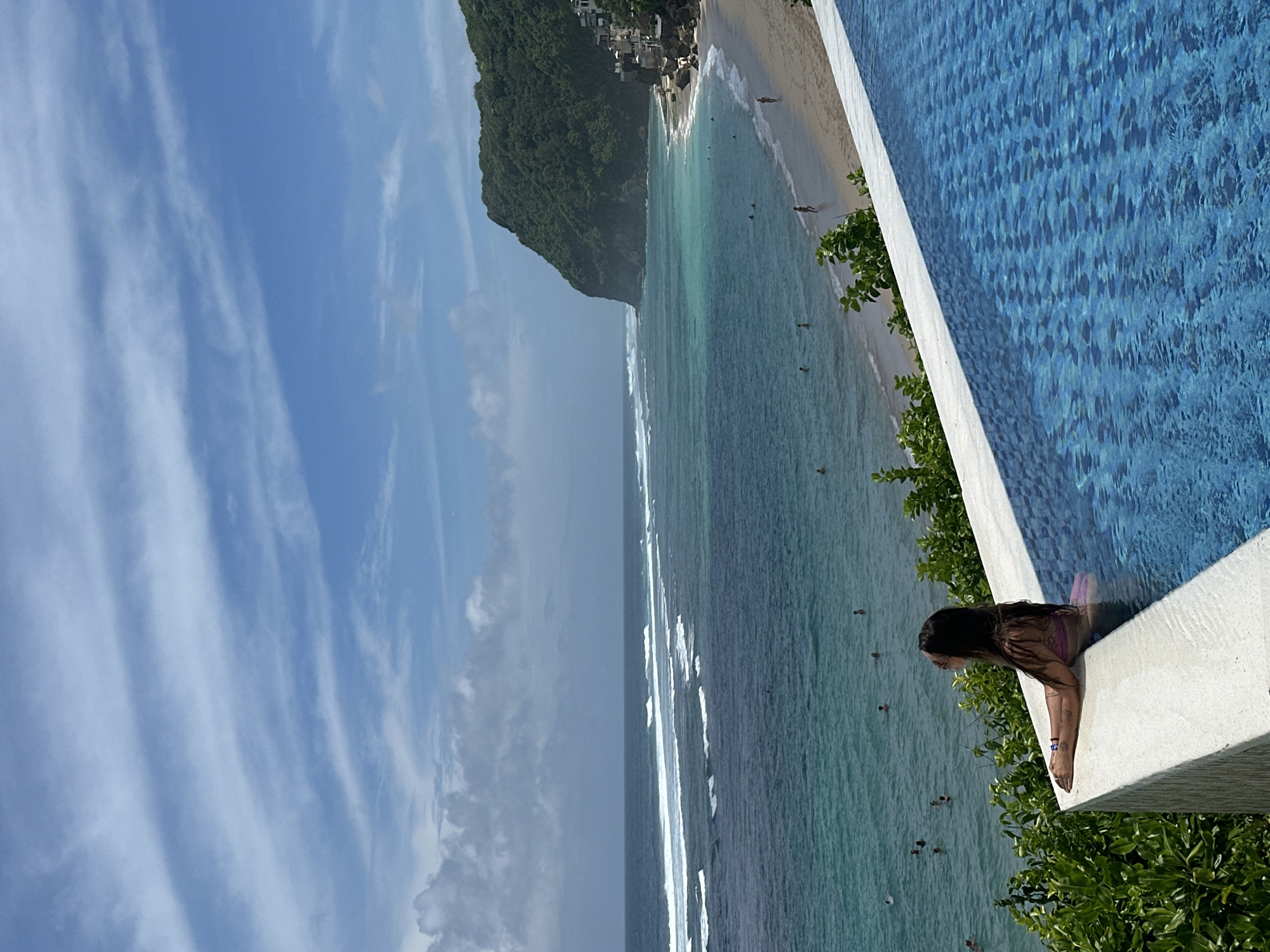 A vibrant tropical coastal scene is captured from an elevated perspective, featuring an infinity pool overlooking a beach and ocean. In the foreground, a person with long dark hair, likely a woman, is seen from the chest up, leaning over the white concrete edge of a blue-tiled infinity pool. She is looking out towards the panoramic view.
Beyond the pool, the turquoise and deep blue waters of the ocean stretch out, with several white-capped waves breaking towards a light sandy beach. Numerous people are visible in the ocean, engaging in activities like swimming or wading, and a few individuals can be seen walking on the beach.
To the left, a large, steep cliff or hill, densely covered in lush green tropical vegetation, dominates the midground. Some buildings are nestled into the landscape at the base and top of this cliff. The sky above is a clear, bright blue with scattered, wispy white clouds, indicating a sunny day. Green foliage from trees and bushes frames parts of the pool area, enhancing the tropical ambiance. The scene depicts a leisurely daytime setting at what appears to be a resort or viewpoint. No text is visible in the image.