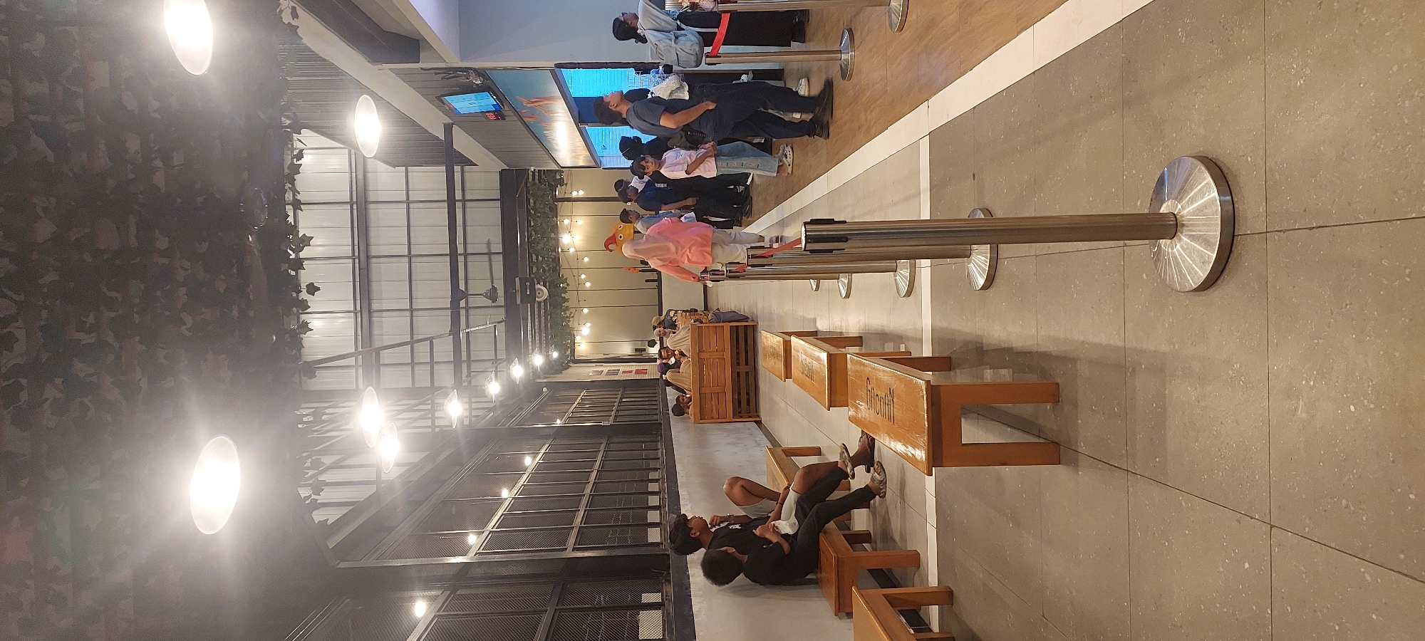 This is a vertical, full-shot photograph taken from a low angle inside a contemporary, dimly lit establishment. The ceiling features a decorative element resembling foliage, and overhead lighting fixtures are visible. The scene depicts a queue of people, with a focus on the individuals lining up.
In the foreground, several wooden benches are arranged, with some occupied by people. Two young males in black t-shirts are seated on a bench to the left, with one looking away from the camera. Further down the row of benches, several more are visible, some empty and some with indistinct figures.
The middle ground shows a longer queue of people, primarily of Asian ethnicity, dressed in casual attire. A woman in a pink hijab and a light-colored top is walking away from the camera, entering the queue. Several individuals are standing, waiting, and observing. The queue is delineated by stanchions with red belts. Behind the queue, wooden seating booths are visible, with some people seated within.
The background reveals more of the establishment's architecture, including a large window with a grid pattern on the left and a wall with a display screen and what appears to be a service counter on the right. The overall environment suggests a public space, possibly a restaurant, cafe, or waiting area.
The image is likely taken in Jayapura, Indonesia, given the context provided. The lighting is somewhat soft and warm, creating a relaxed ambiance. The floor is tiled with a speckled gray material.