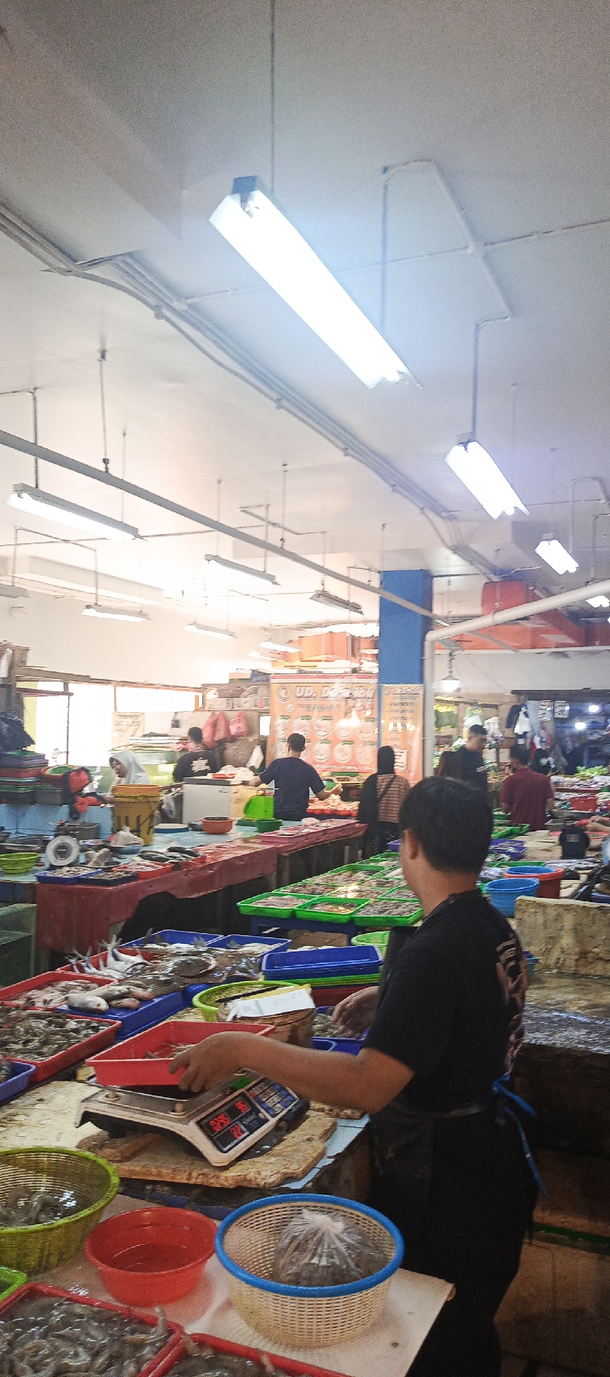 The image depicts a busy indoor market scene, likely a fish market, in Sukaresmi, Indonesia. The ceiling is illuminated by multiple fluorescent tube lights, with visible conduits running across it. In the foreground, a person wearing a black t-shirt and apron is operating a digital scale placed on a butcher block. Various containers of seafood, including shrimp and fish, are arranged on tables and in baskets around the scale. In the background, other individuals are seen attending to their stalls and customers. One person is wearing a headscarf, while others are dressed in casual attire. Several signs with text and images are visible on the stalls, suggesting a commercial setting. The market appears well-stocked with fresh seafood.