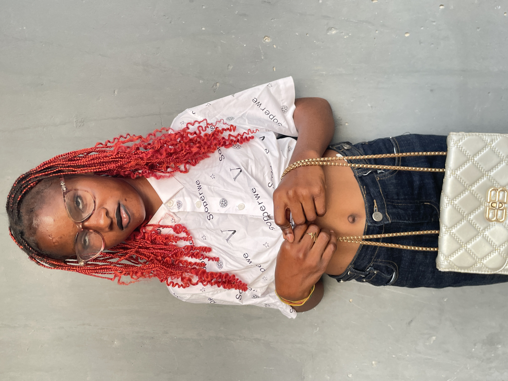 A young person, likely female, with dark skin and distinctive bright red curly braids, is captured in a portrait while lying on a light grey, possibly concrete, surface. They are wearing clear-framed eyeglasses and gazing directly at the camera with a calm expression.
The individual is dressed in a white collared, button-down shirt patterned with various black designs including stars, geometric shapes, and repeating text such as "Soperwe" and "Sor." They are also wearing dark blue denim jeans. Adorning their hands and neck are several gold-colored accessories, including a chain necklace, a bracelet, and rings, one of which features a distinctive "W" or heartbeat-like design. A light-colored, possibly silver or off-white, quilted shoulder bag with a prominent gold "BB" logo and a gold chain strap rests beside them.
The setting is simple, suggesting an outdoor or semi-outdoor location with a plain, textured ground surface. The overall impression is a casual, posed photograph, set in Badagry, Nigeria.