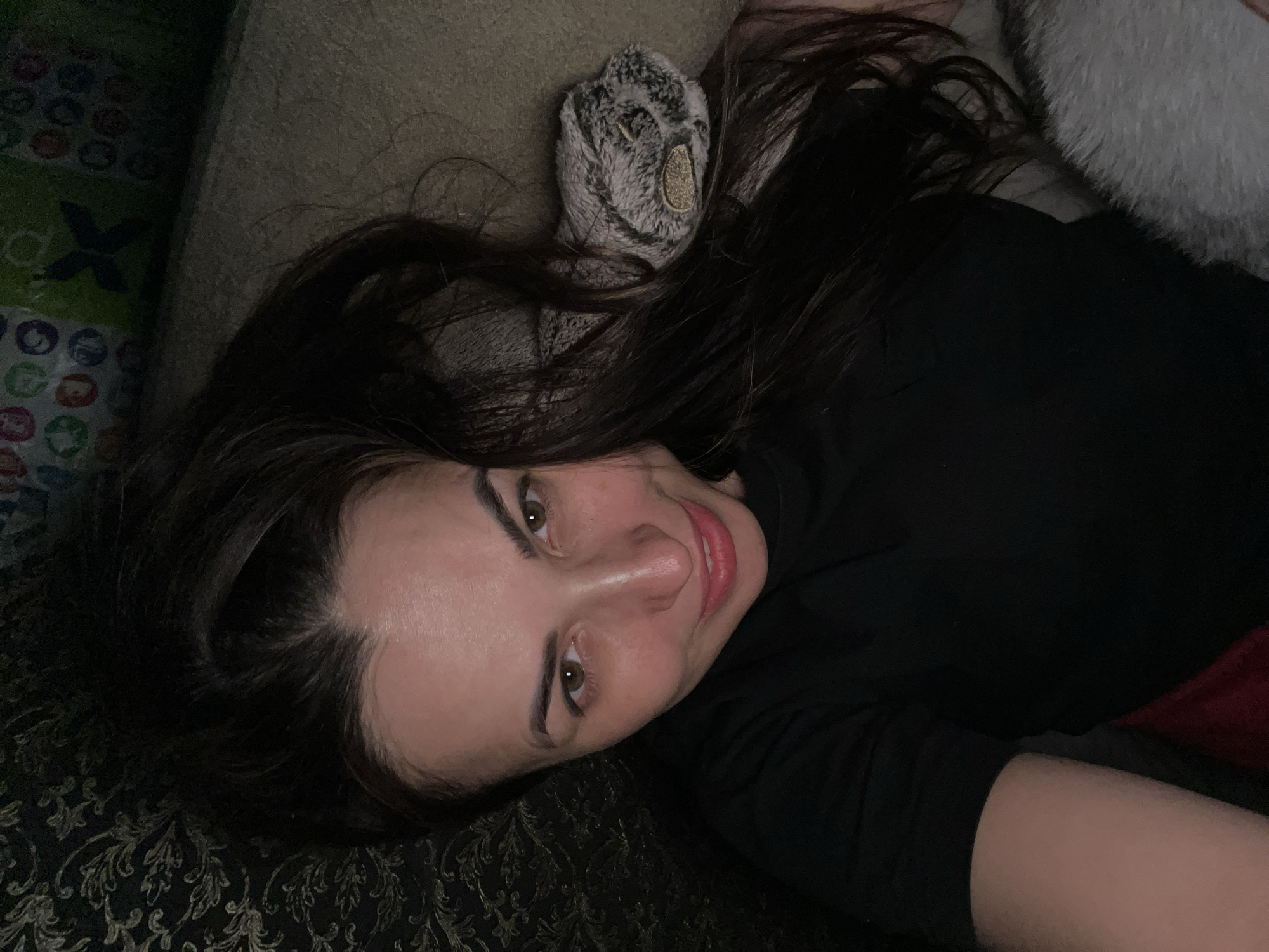 A woman lies in a supine position. Her head rests on a patterned surface, and her hair is spread around her. She wears a dark top. Adjacent to her head are a cushion or pillow with a patterned cover and a fluffy, light-colored object.
