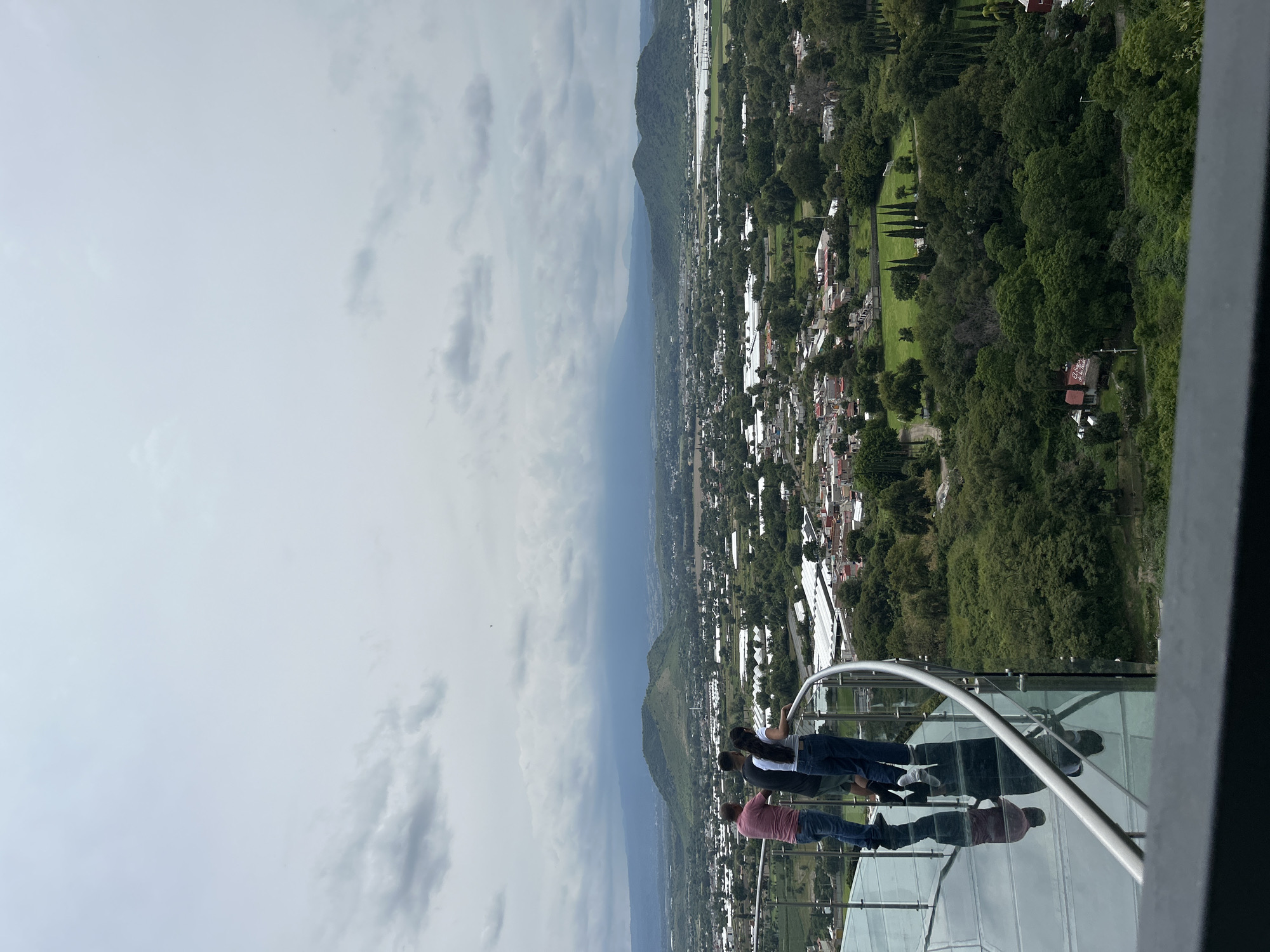 The image shows a panoramic view from a glass-floored observation deck in Atlixco, Mexico. Three individuals are standing on the deck, looking out at the landscape. The deck has a curved railing made of stainless steel. Below the deck, the glass floor reflects the sky and the people. The view includes a sprawling city with buildings and greenhouses, surrounded by lush green trees and rolling hills. In the distance, mountains rise under a cloudy sky. The overall impression is one of a scenic overlook providing a breathtaking perspective of the city and its natural surroundings.