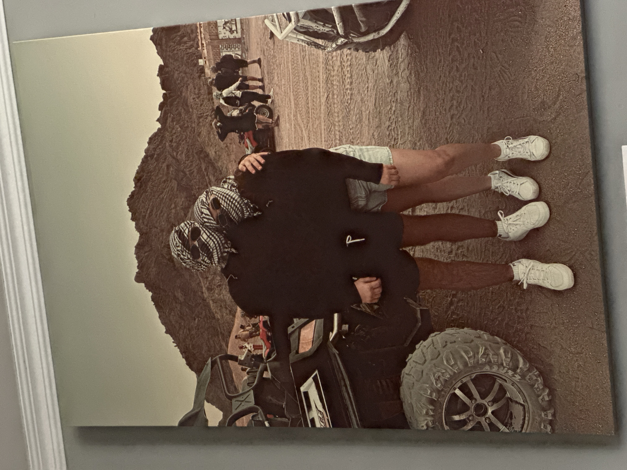 A candid, medium shot captures two individuals embracing, their faces concealed by checkered keffiyeh scarves. The person on the left wears sunglasses, while the person on the right also has sunglasses visible beneath their scarf. They are positioned in front of a black ATV, with a large tire prominent in the foreground. Both individuals are dressed in casual attire, with the person on the left wearing shorts and the person on the right wearing denim shorts. They are standing on a sandy surface in a desert landscape, with rugged mountains in the background. Several other ATVs and people are visible in the middle ground, suggesting an activity or tour. The lighting is natural, with a slightly muted color palette. The overall scene depicts a casual outing in a desert environment.
