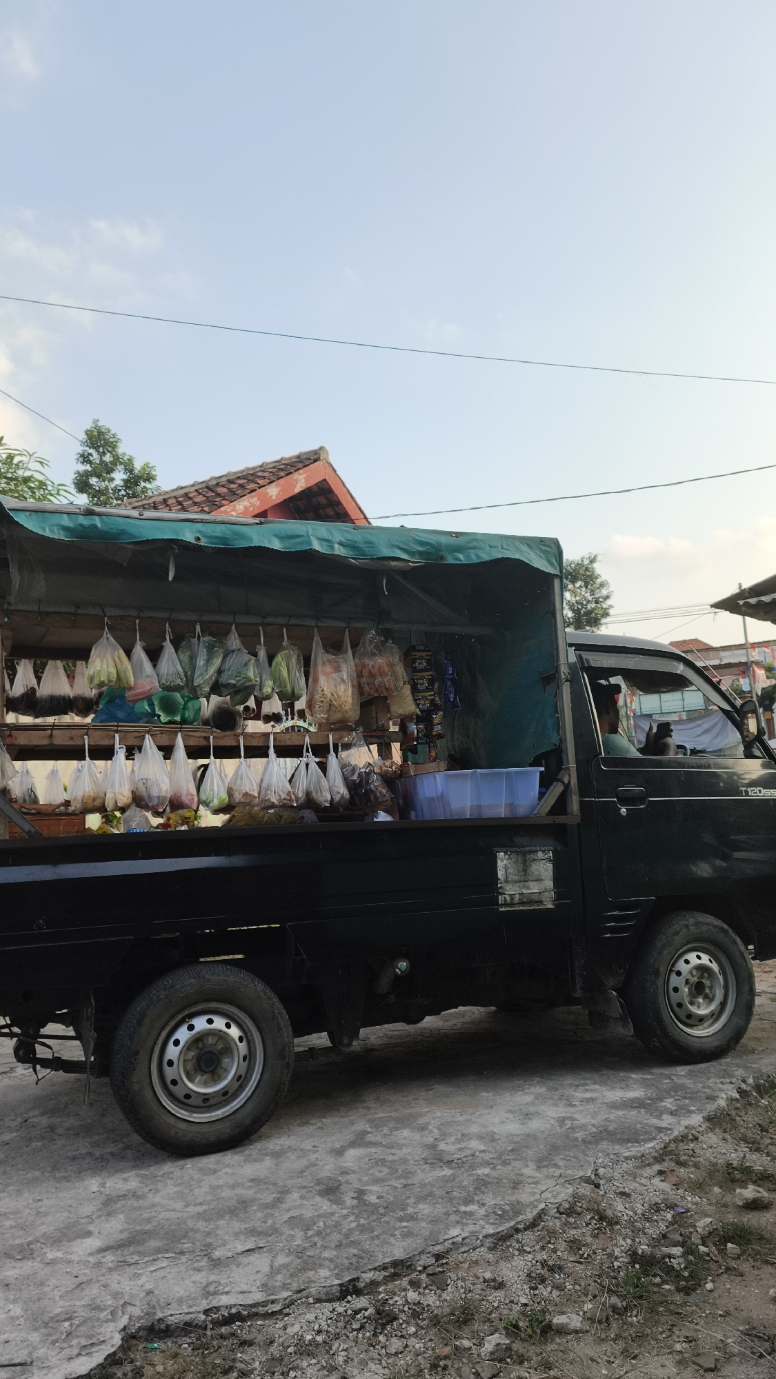 An outdoor, slightly low-angle, full shot shows a dark green pickup truck parked on a concrete surface. The truck's bed is converted into a makeshift store, covered by a green tarp and displaying an assortment of goods. These goods are primarily packaged in plastic bags and hung from a horizontal bar, with some items also placed on shelves within the truck bed. The items appear to be a mix of produce and packaged food products. A person is visible inside the driver's seat of the truck, holding a phone. The background features a red-tiled roof of a building and some trees. The sky above is a pale blue with a few wispy clouds, suggesting it is either early morning or late afternoon. The overall scene depicts a mobile market or roadside vendor in what appears to be a semi-urban or rural setting in Bandar Lampung, Indonesia.