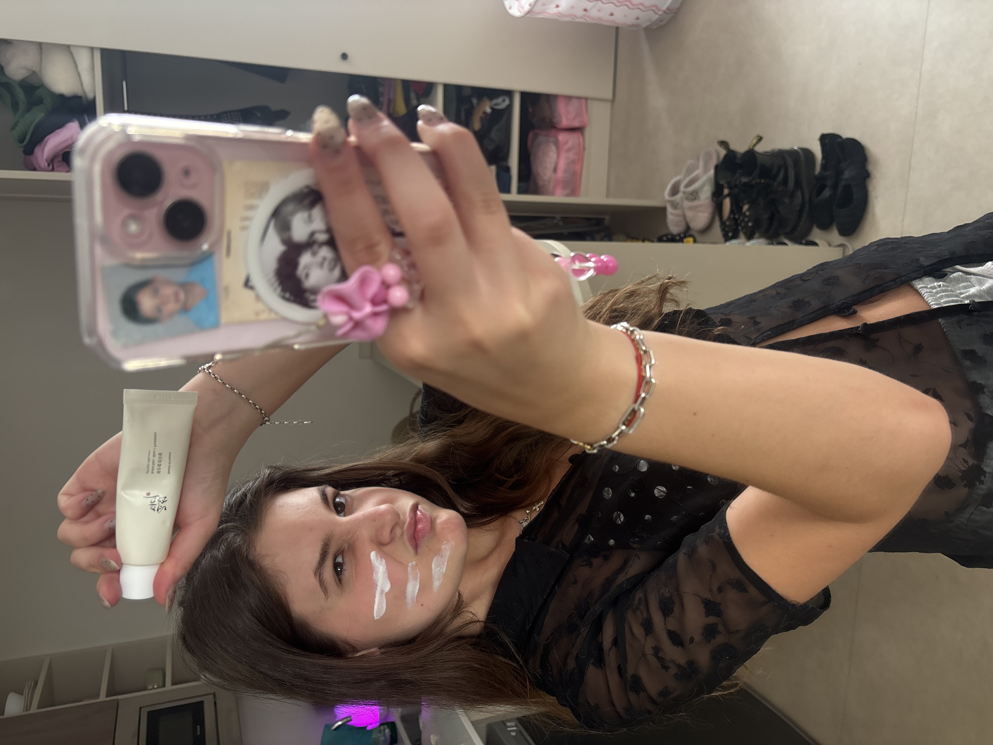 A young woman is taking a selfie in what appears to be a bedroom or dressing area. She holds a white tube of cream in her left hand and has applied three streaks of white cream to her face. She is wearing a black sheer top. She is holding a phone with a decorated case, including a pink flower and stickers. Behind her, there is a wardrobe with clothes visible inside, and a shelf with shoes. The exact location within Getafe, Spain cannot be determined from the image.