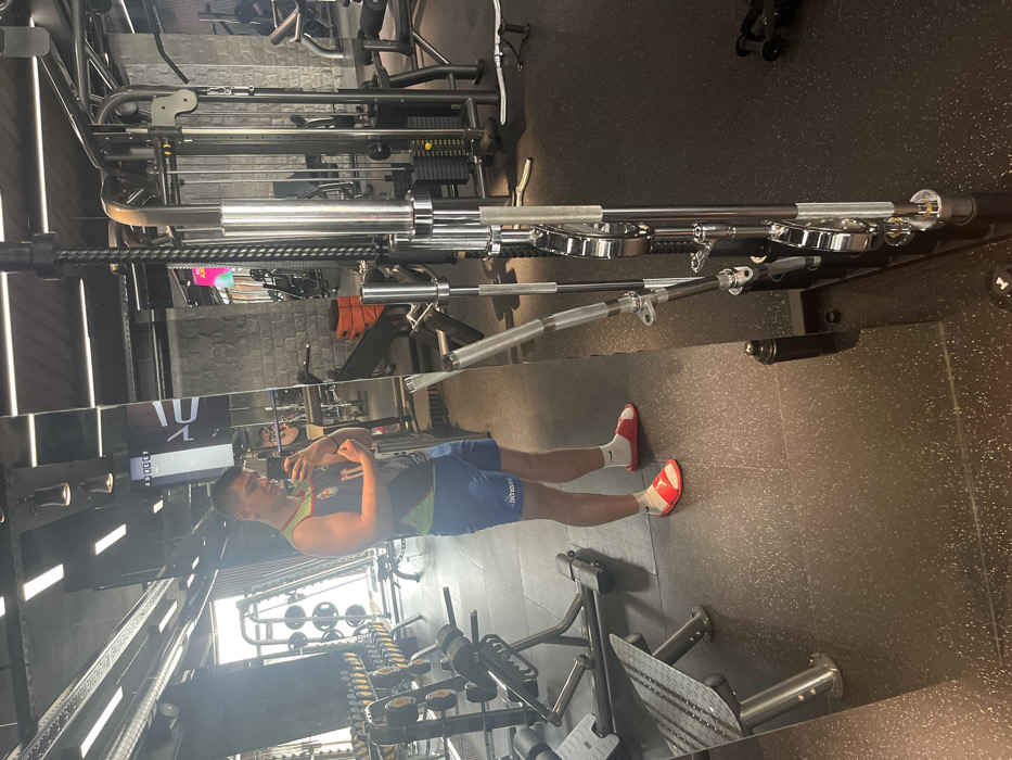 An adult male stands in a fitness center in Sofia, Bulgaria, posing for a mirror selfie. He is observed from the waist up in the direct reflection and full body in the mirror panel to his left. The male is wearing a dark sleeveless athletic top with a multi-colored emblem on the chest, blue athletic shorts displaying "SKYSPORT" text, white socks, and red slide sandals. He holds a smartphone in his left hand at chest height, pointed towards himself, while flexing his right bicep. The gym environment features dark, speckled rubber flooring and large mirrored wall panels. In the background, various pieces of gym equipment are visible, including a dumbbell rack, weight benches, multiple cable machines, and a barbell storage rack with chrome barbells. Overhead lighting consists of rectangular LED panels and spotlights. A black shaker bottle is present on the floor to the right of the subject's feet.