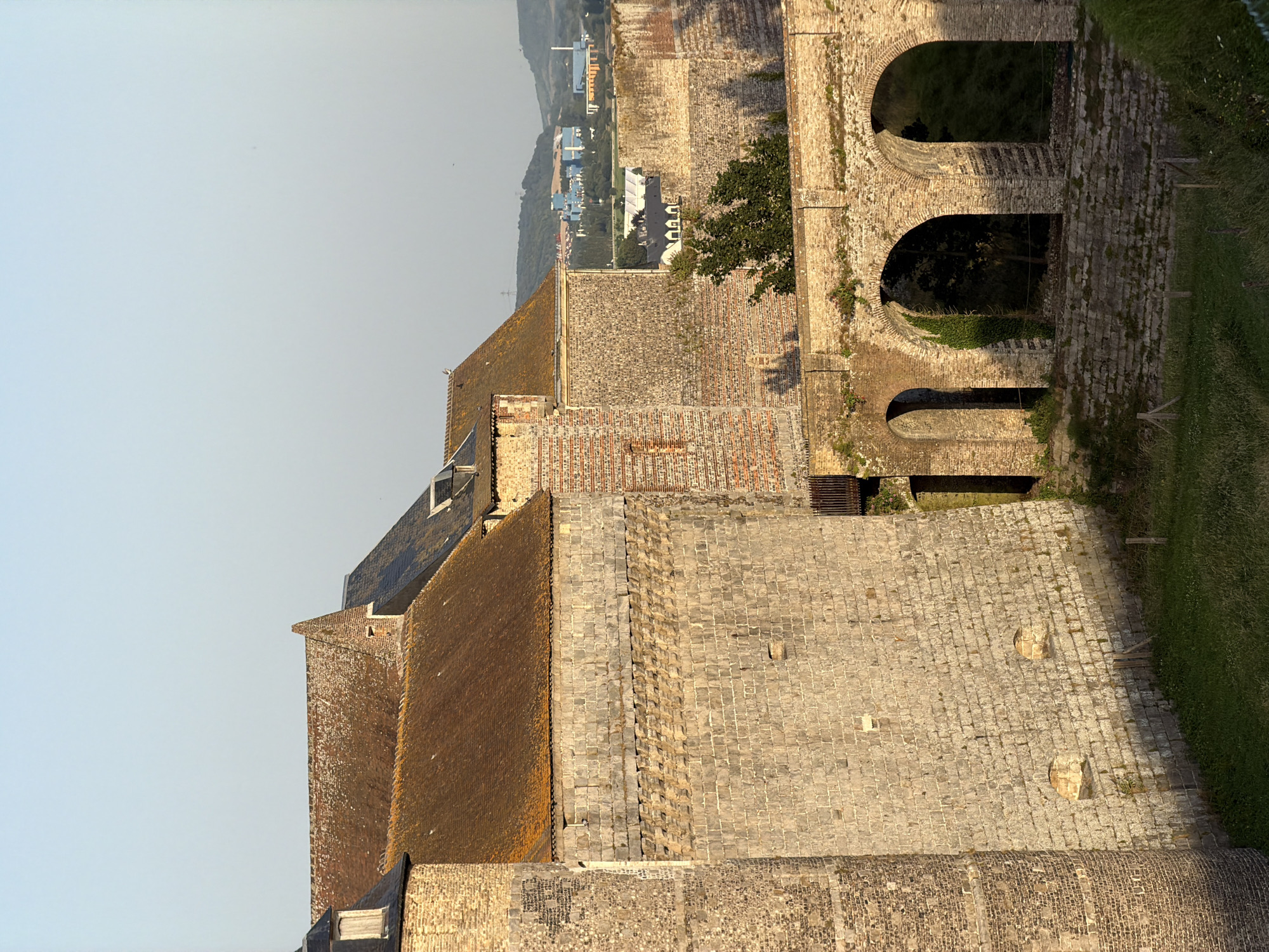 This image depicts a historical stone structure, possibly a fortress or castle, in Dieppe, France. The perspective is from an elevated vantage point, looking down and across the extensive stone buildings and their tiled roofs. The stone masonry is varied, with larger, rough-hewn blocks forming the main walls and smaller, more uniform stones used for detailing and pathways. Several arched openings are visible in one section of the building, suggesting an older architectural style.
The scene is bathed in natural light, indicating it was taken during the daytime, likely late afternoon or early morning given the warm, golden tones on the stone. The sky is clear and pale blue, suggesting fair weather. Beyond the immediate structures, a distant landscape unfolds, featuring rolling hills with scattered buildings and trees, hinting at the surrounding town or countryside. A patch of green, well-maintained grass is visible to the right, possibly a lawn or park area adjacent to the fortress.
No people are visible in the image. The focus is entirely on the architecture and the vastness of the historical complex. The overall impression is one of enduring history and solid construction, with a sense of peace and quietude due to the absence of human activity. The weathered appearance of the stone and tiles suggests the age of the structure.