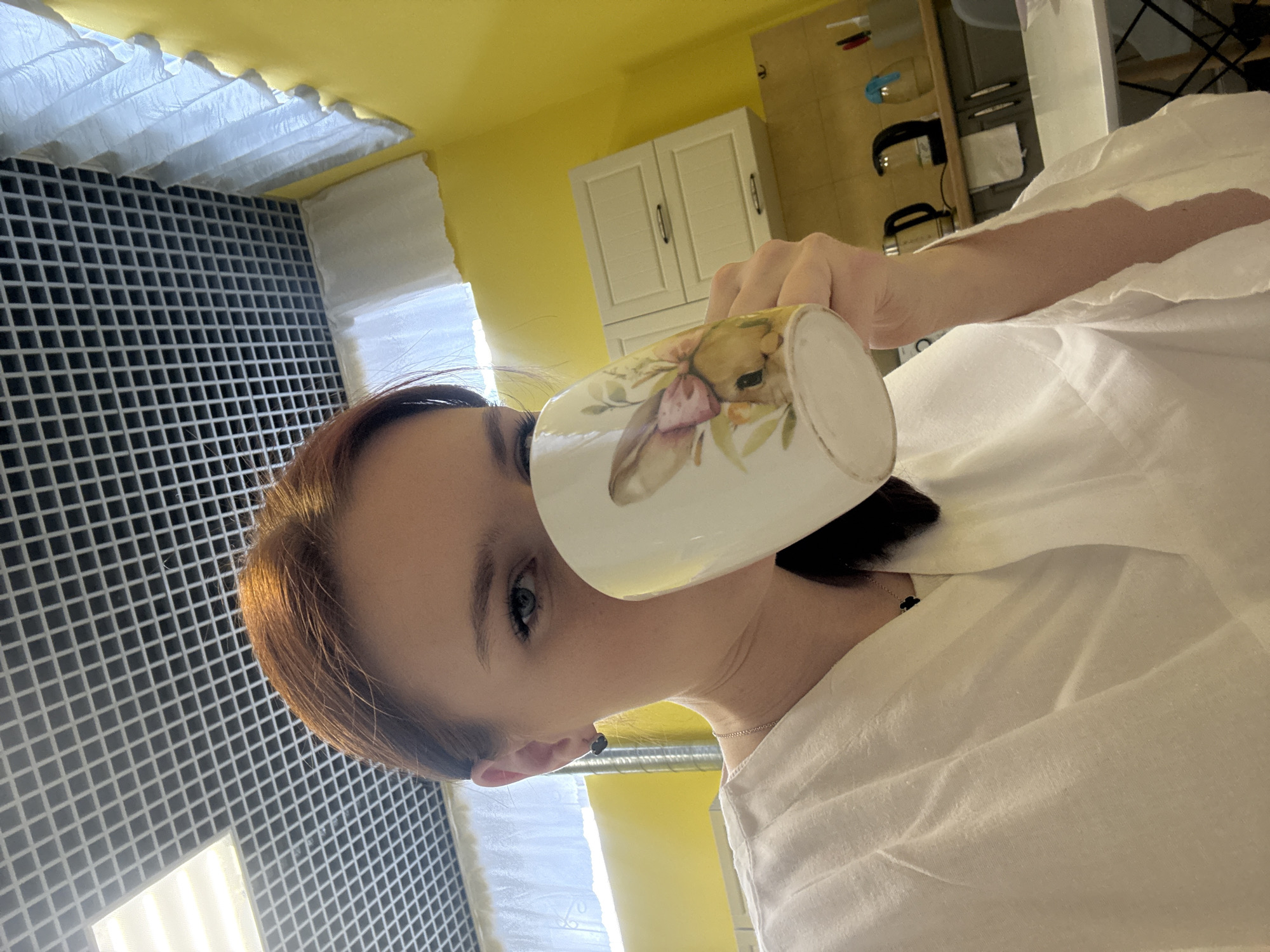 Here is a detailed description of the image:
A woman is holding a ceramic mug with a bunny and floral design. She is wearing a white shirt or robe and has auburn hair. A clover-shaped earring is visible on her left ear, and a delicate silver necklace adorns her neck. The background reveals a room with yellow walls, white window treatments, and a grid-like patterned structure, possibly a window covering. In the upper portion of the frame, kitchen cabinets and utensils suggest this is a kitchen environment.
