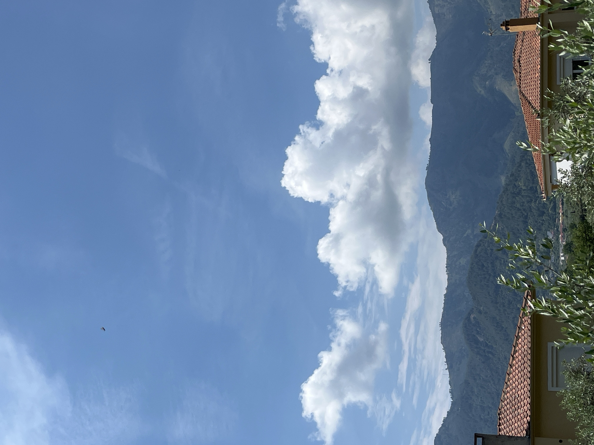 An outdoor, low-angle, medium shot captures a view of rolling green hills under a bright blue sky dotted with fluffy white clouds. In the distance, a tiny, dark object, possibly a bird or aircraft, is visible in the upper left quadrant of the frame. The foreground features the terracotta tiled roofs of two houses, partially obscured by lush green foliage in the lower half of the image. One house on the left shows a sliver of its yellow exterior wall. The second house on the right has a prominent chimney. The overall scene suggests a sunny day in a semi-rural or suburban setting with a mountainous backdrop. The location is Golden Beach, Greece.