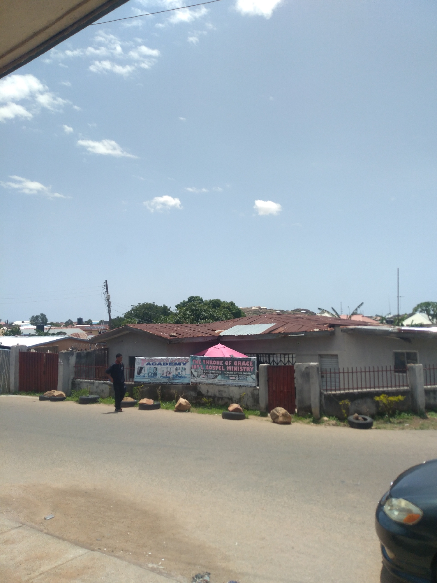 This is a street-level, eye-level, medium-shot photograph taken on a sunny day in Jos, Nigeria. The scene depicts a street with a building facade and a sidewalk in the foreground. A dark-colored sedan is partially visible on the right side of the frame, parked on the street. A lone individual, appearing to be a man in a dark uniform, stands facing the street in front of a building. This building features a large banner advertising "PRELUDE STONE ACADEMY" and "THE THRONE OF GRACE INT'L GOSPEL MINISTRY". The banner also indicates offerings such as "DAYCARE, NURSERY, PRIMARY, SECONDARY" and "SCHOOL OF THE SWORD". Behind the building, a cluster of houses with corrugated iron roofs is visible. The sky above is a clear blue, dotted with scattered white clouds. The street surface is asphalt, and the sidewalk appears to be concrete. Several tires and rocks are strategically placed along the edge of the sidewalk and in front of the building.