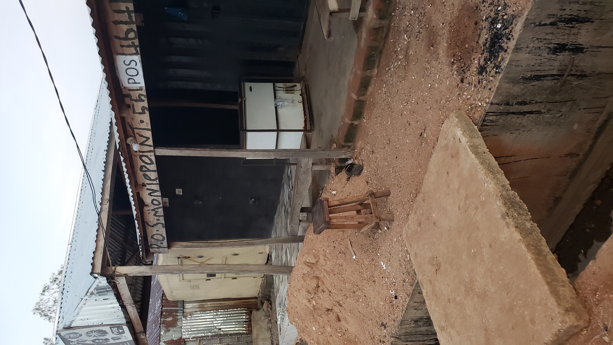 The image shows an outdoor scene in Jalingo, Nigeria, featuring a weathered structure that appears to be a small shop or kiosk. The building has a corrugated metal roof supported by wooden posts. Painted on the wooden beam above the shop's entrance is lettering that reads "P.O.S. MONIEPOINT 561 POS 464". In front of the structure, there is a pile of reddish-brown dirt and gravel. A simple wooden stool sits on the ground. A white chest freezer with two doors is visible inside the shop. Further in the foreground, a concrete slab bridges a dark, narrow trench or channel, with the slab showing signs of wear and possibly being part of a makeshift walkway or bridge. The surrounding environment is rough and dusty, with some scattered debris. The overall impression is of a modest, functional establishment in a developing area.
