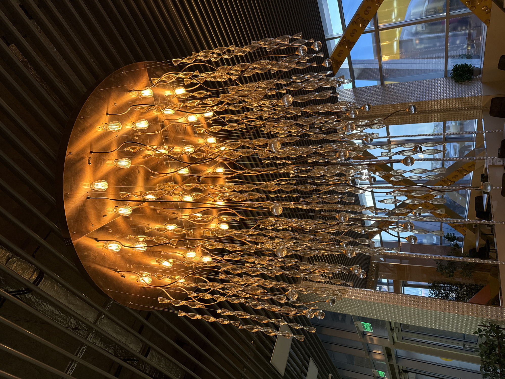 The image depicts a large, ornate chandelier hanging from a ceiling. The fixture has a circular base with numerous exposed light bulbs. Hanging from the base are many strands of clear, twisted glass or crystal pieces that appear to resemble elongated fish. The background includes glimpses of a modern building interior with large windows and architectural elements. There are reflections of the interior in the windows. The ceiling above the chandelier has a corrugated metal appearance. The location in the city of Petropavlovsk-Kamchatsky, Russia, cannot be determined from the visual evidence.