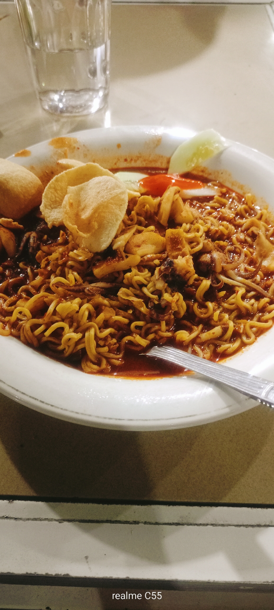 A close-up shot showcases a plate of mie goreng, a popular Indonesian stir-fried noodle dish, served in a white bowl. The noodles are coated in a rich, dark sauce, interspersed with pieces of squid and possibly other seafood. Two round, light-colored crackers, likely kerupuk, are placed on top of the noodles. A slice of cucumber and a dollop of red chili sauce are also visible on the side, suggesting a spicy accompaniment. A fork rests on the edge of the bowl, indicating that the meal is ready to be eaten. In the background, a glass filled with a clear liquid, possibly water, and a blurred table surface are visible. The lighting suggests an indoor setting, possibly a restaurant, and the time of day is unclear. The overall impression is of a delicious and hearty meal. The text "realme C55" is visible at the bottom of the image. This image was taken in Lhokseumawe, Indonesia.
