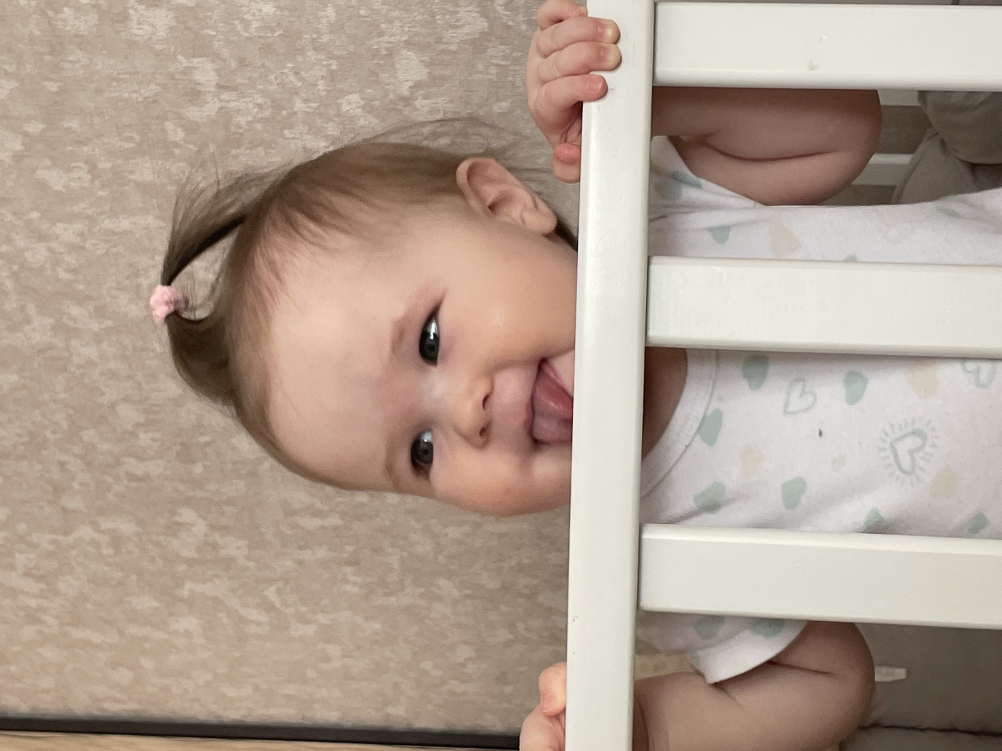 A baby with a small ponytail secured with a pink hair tie is standing in a white crib. The baby is sticking their tongue out and looking towards the camera. The crib is positioned against a textured beige wall. The location is Chaikovsky, Russia.