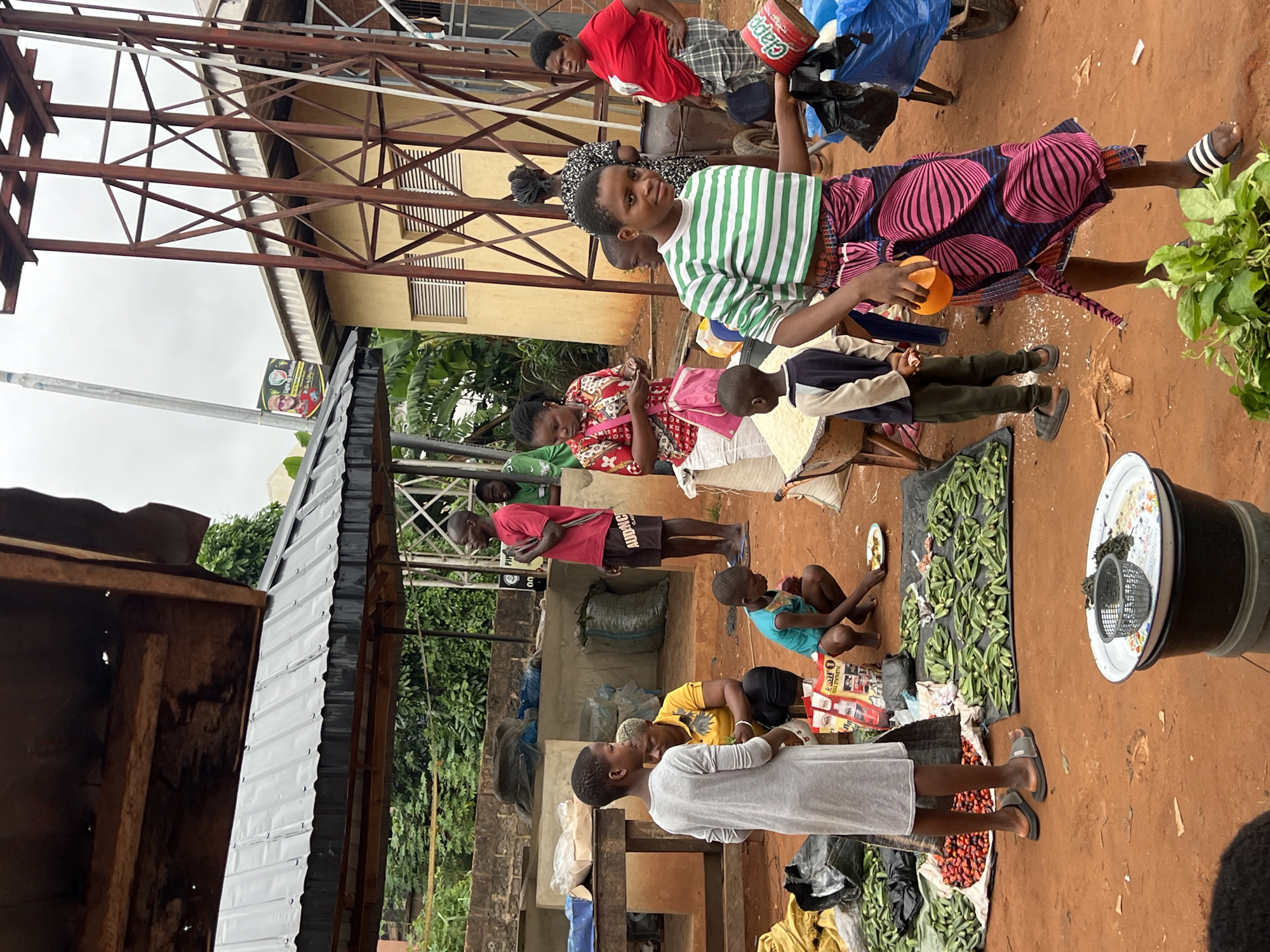 This is a photograph taken at an outdoor market in Nkpor, Nigeria. The scene depicts a bustling market with various individuals engaged in commerce. A young girl in a gray dress stands in the foreground, looking to her left. To her right, a woman in a yellow and black patterned shirt kneels, holding some items. A young boy in a teal top is also crouched down, eating from a plate.
In the background, several other people are visible. A man in a red shirt and dark shorts stands facing left, holding his hand to his chest. Other individuals are seen in various poses, suggesting activity within the market. The market stalls are constructed with corrugated metal roofs and wooden supports. Produce, including what appears to be okra and red berries, is spread out on black tarps on the ground, indicating fresh goods for sale. Bags of what might be grain or other commodities are also present.
The environment appears to be an urban or semi-urban setting, with buildings and metallic structures visible in the background. A poster is attached to a pole in the distance. The ground is covered in reddish-brown earth. The overall atmosphere suggests a typical day at a lively Nigerian market.