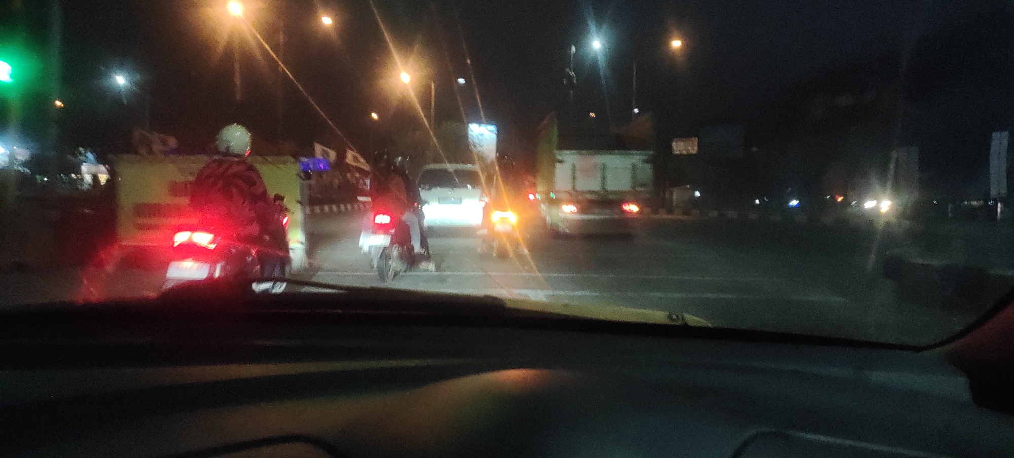 The image depicts a nighttime scene from inside a vehicle, looking out at traffic on a road in Mranak, Indonesia. The foreground shows the dashboard of the vehicle. The middle ground is dominated by a line of vehicles, including motorcycles and a white van. One motorcycle in the center has its brake lights illuminated. A rider on a motorcycle with a red jacket is visible on the left. A large truck with red taillights is seen further down the road. Streetlights and vehicle headlights create a bright, somewhat blurred effect in the dark environment. The road appears to be paved and there are indications of street infrastructure and signage in the background, including what appear to be billboards. The overall impression is one of a moderately busy urban or suburban road at night.