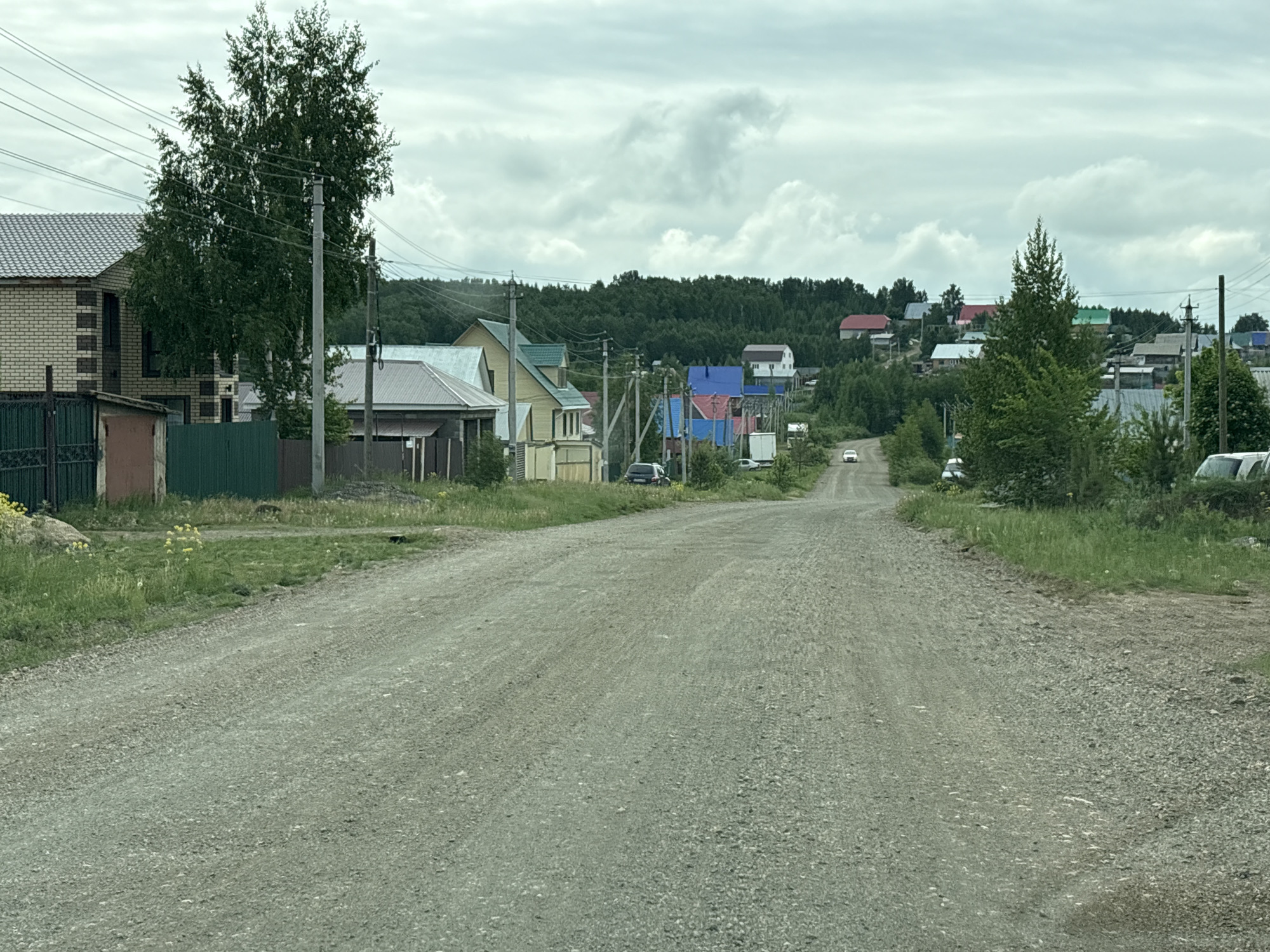 The image depicts a gravel road in a rural or suburban setting, likely within the city of Unknown, Unknown. The road extends into the distance, flanked by houses and greenery on either side. Houses with diverse roofing colors, including red, blue, and green, are visible. Power lines run above the road. A forest area is visible in the background on a slight incline. Several vehicles are parked on the sides of the road, and one white car is further down the road. The sky is overcast with cloudy weather conditions.
