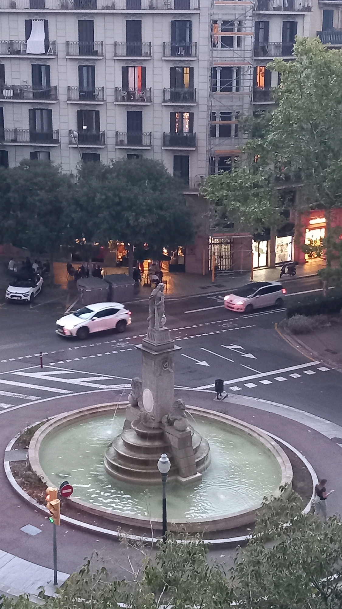 The image shows a street scene in Barcelona, Spain, likely in the Eixample district given the characteristic apartment buildings with balconies and mansard roofs. In the foreground, a large, multi-tiered fountain with sculpted lions is the central focus. Water is flowing from the fountain into the circular pool. A traffic light with a green signal is visible to the left of the fountain.
A white SUV is driving on the circular street around the fountain. To the left of the fountain, a white convertible car is parked. In the background, a multi-story apartment building with numerous balconies and windows is visible. Some windows are illuminated, suggesting occupancy. Several trees with lush green foliage are scattered throughout the scene. There are also shops with illuminated storefronts and outdoor seating areas with people seated at tables. A person is walking near the fountain on the right side of the image. A scooter is parked outside one of the shops.