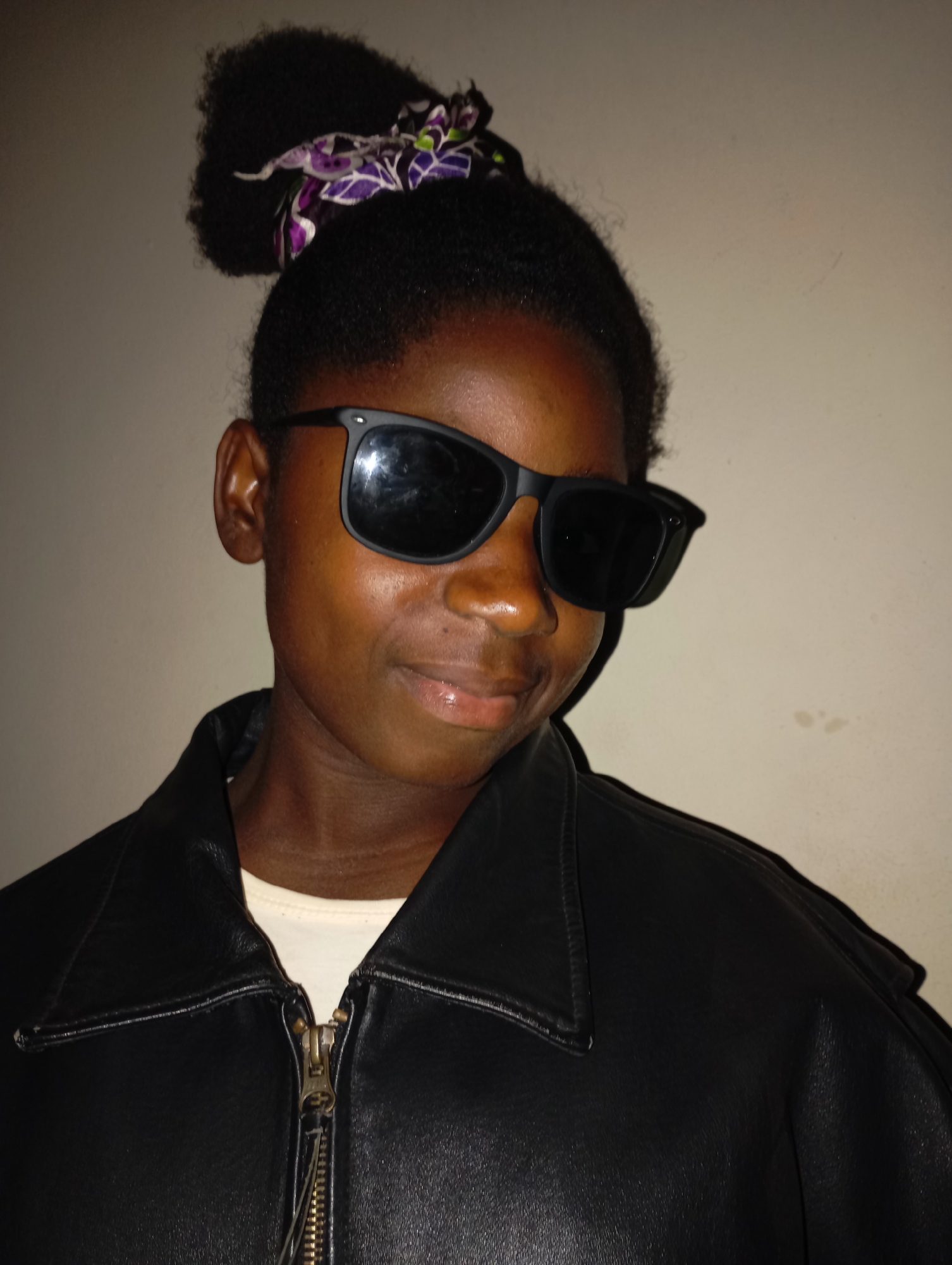 A medium shot depicts a person wearing a black leather jacket and sunglasses. The person has dark skin and their hair is styled in a high bun with a patterned hair accessory. The background is a plain, off-white wall. There is no visual information to confirm the location is in Jos, Nigeria.