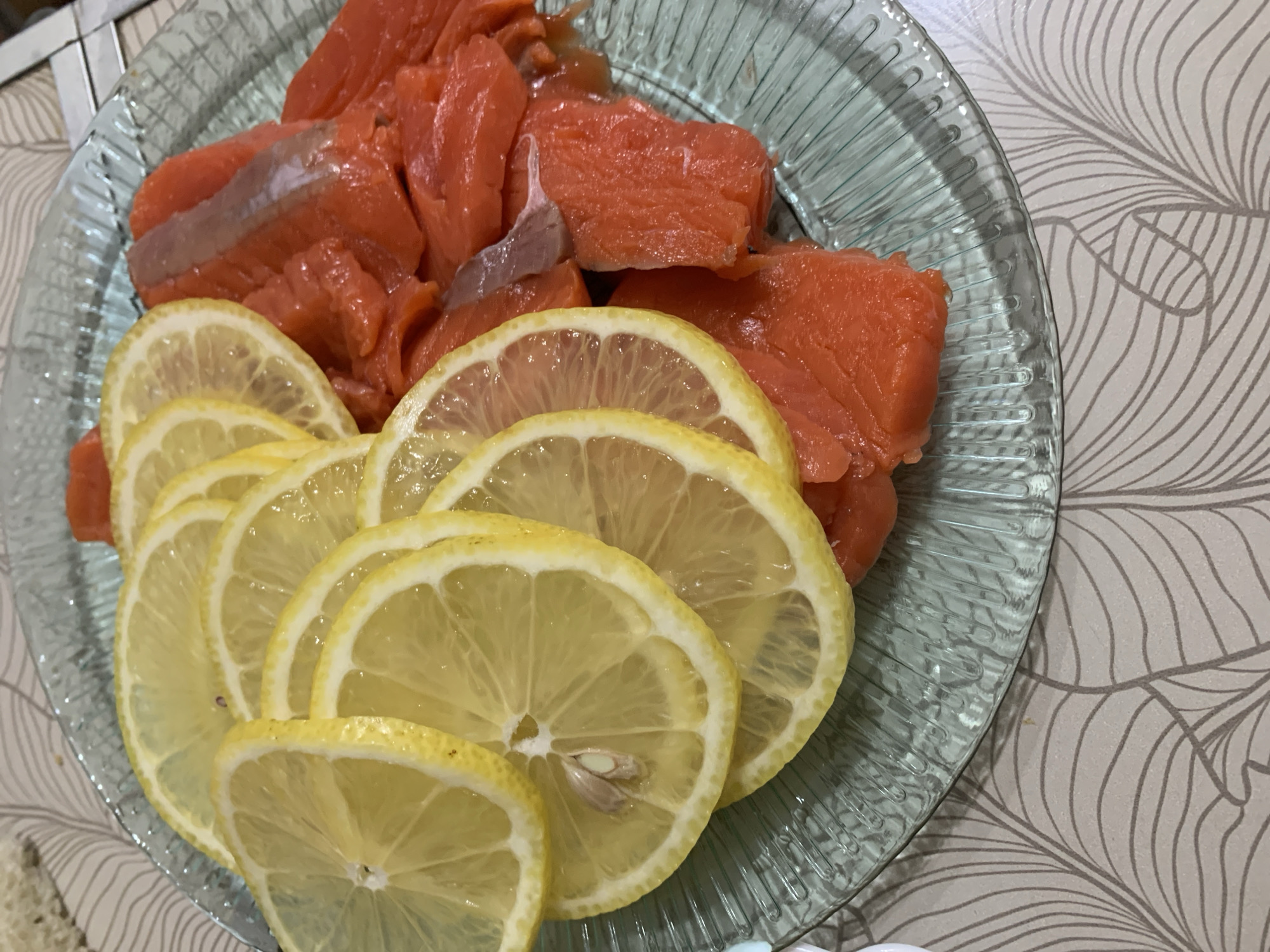 A close-up shot shows sliced salmon and lemon on a decorative glass plate. The salmon is arranged on the right side of the plate, appearing as vibrant orange-red pieces. Thin slices of lemon are fanned out on the left side of the plate, some overlapping. A single lemon seed is visible in the center of one of the lemon slices. The plate has a ribbed pattern and rests on a surface with a light-colored, patterned tablecloth featuring abstract leaf-like designs. The lighting is somewhat dim, casting soft shadows. This image was taken in Magnitogorsk, Russia.