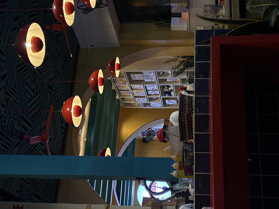 A vibrant interior space features distinctive red and white globe pendant lights suspended against a dark, patterned wall. A teal beam crosses the foreground, leading the eye towards a counter area with condiments and a glimpse of staff. In the background, decorative shelves display items, and an arched opening reveals a fan and a bright, circular sign with "UCHO" visible. The scene is a mix of bold colors, geometric shapes, and functional elements.