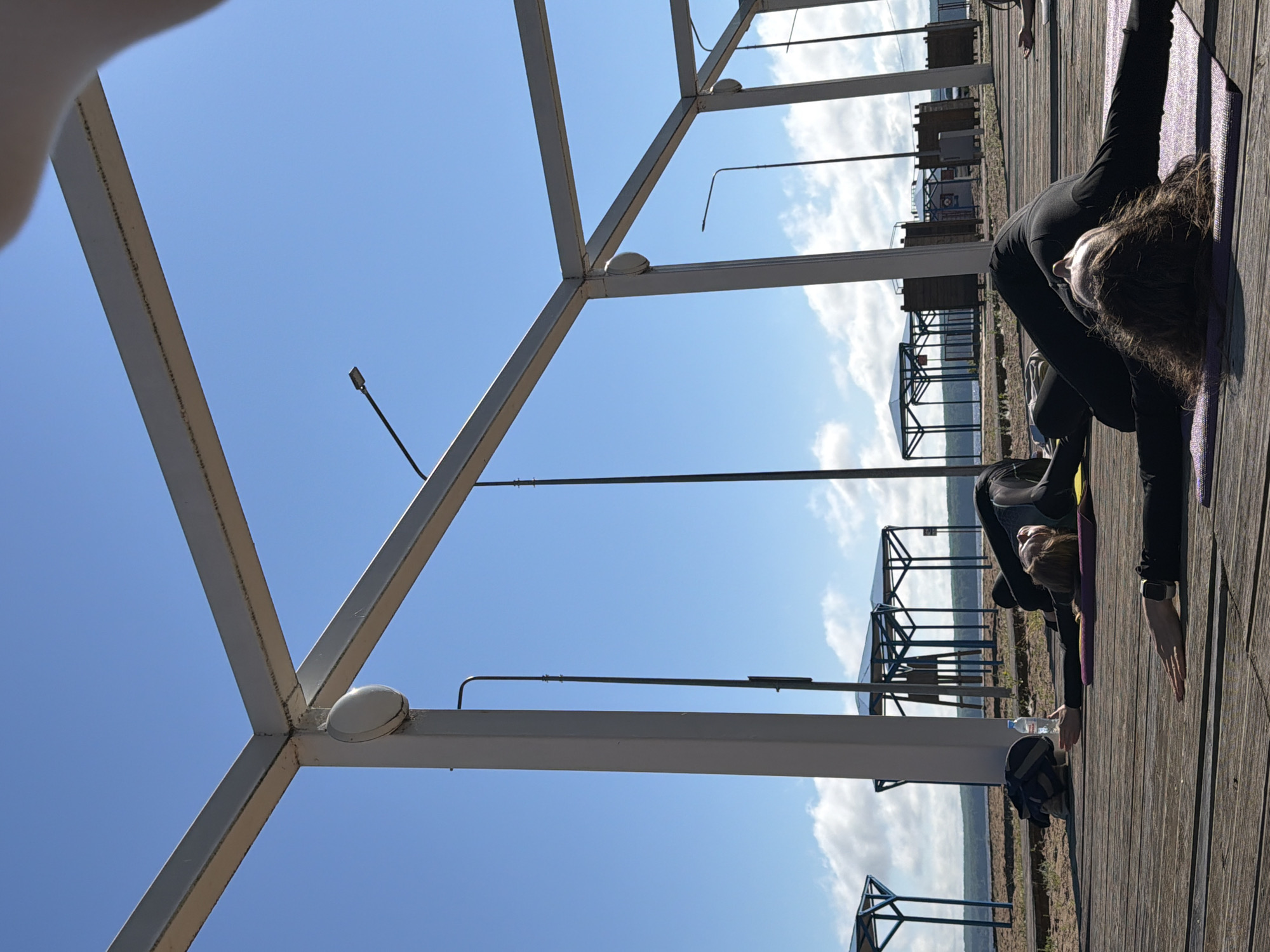 The image depicts an outdoor yoga session on a wooden deck, likely on the embankment of the Kama River in Chaikovsky, Russia. The sky is a clear blue with scattered white clouds. A modern, light-colored metal structure with large beams forms a frame over the scene, suggesting it is part of a public space or recreational area. Several people are visible, dressed in dark athletic wear, lying on yoga mats in a relaxed, supine position. In the background, there are covered shelters or gazebos, beach chairs, and a body of water, indicating a waterfront location. The overall atmosphere is one of peaceful outdoor activity.