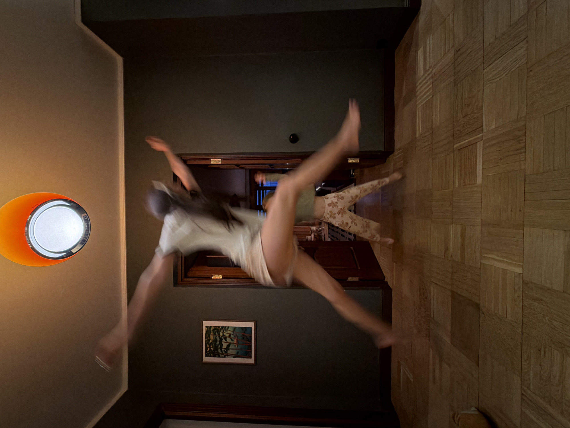 The image shows an interior scene with a blurred motion effect, making details difficult to discern clearly. Two figures are visible in motion, one closer to the foreground wearing a light-colored shirt and shorts, and another farther back wearing a green top and patterned pants. An orange-and-white lamp is attached to the wall on the left. A framed picture hangs on the wall, and a doorway is visible in the background. The floor is made of wood planks laid out in a square pattern. The location is unspecified but said to be in Khimki, Russia.