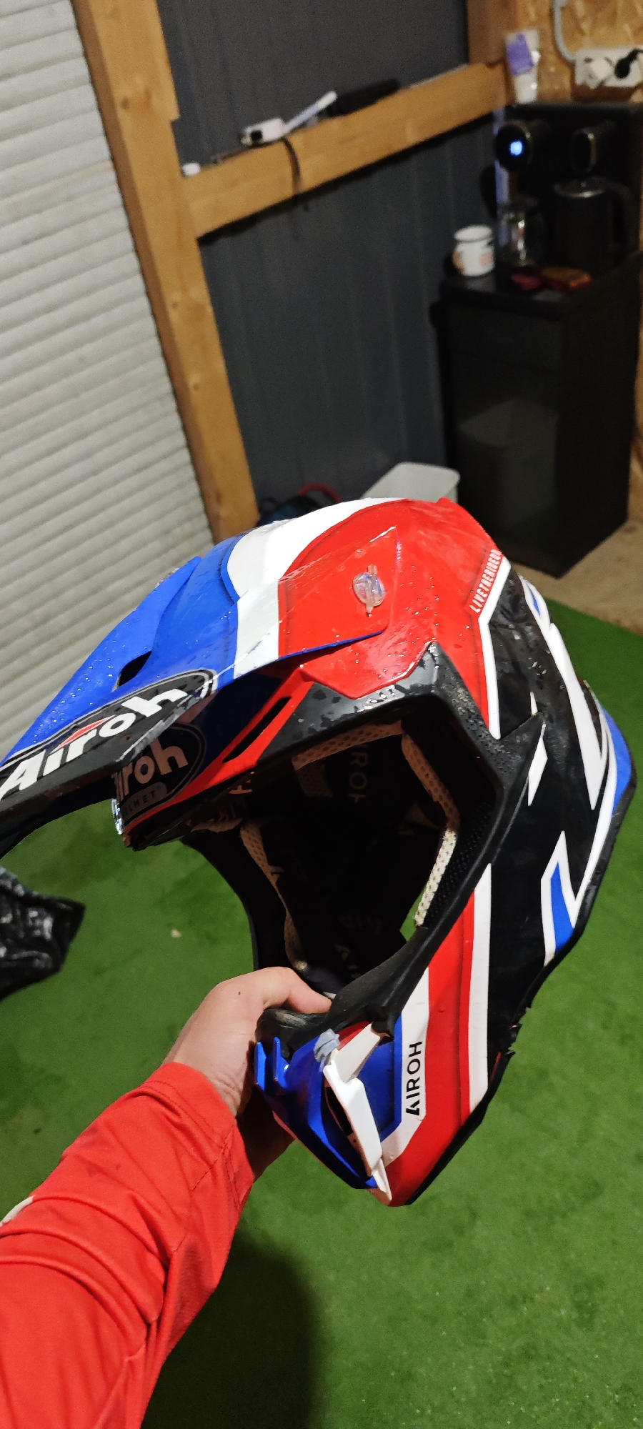 The image shows a person holding a blue, white, and red AIROH dirt bike helmet. The helmet has the AIROH logo prominently displayed. The person is wearing a red long-sleeved shirt. The background includes a green surface, possibly artificial turf, a wooden structure, and a black cabinet with a coffee maker on top. The location is Kazachiy Brod, Russia.