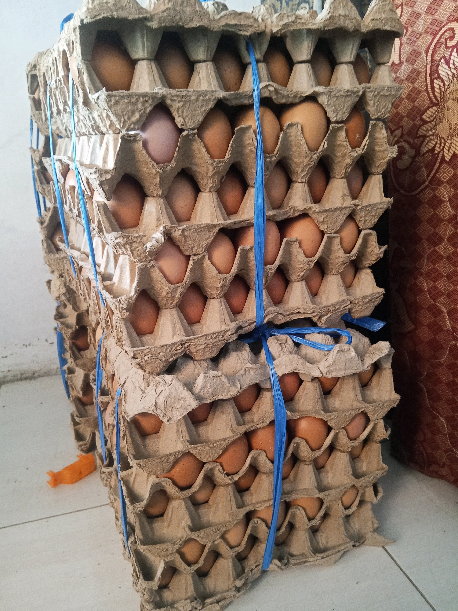 Two stacked towers of cardboard egg cartons, each holding brown eggs, are secured with blue plastic ties. The cartons are positioned on a light-colored tiled floor. To the right of the egg cartons, a textured reddish-brown fabric is visible. In the background, a plain white wall is present. An orange plastic object, possibly a clip or small toy, lies on the floor to the left of the egg cartons. The overall scene appears to be indoors, in a storage area or market stall, consistent with a location in Jinang Kampung Pansur, Indonesia.