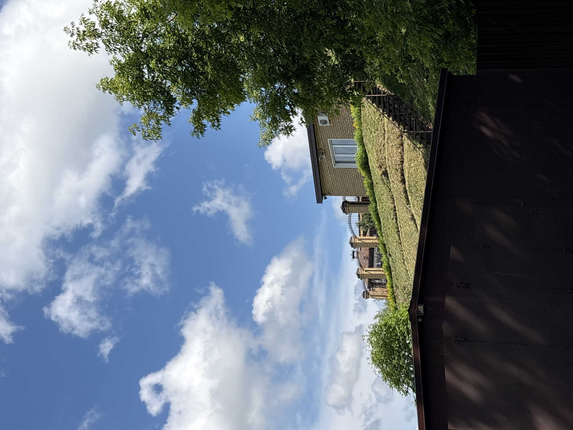 The image depicts a sunny day with a bright blue sky dotted with fluffy white clouds. In the foreground, a large, dark brown, corrugated metal structure, possibly a garage or workshop, occupies the lower portion of the frame. Its surface is partially illuminated by dappled sunlight, creating shadows.
Behind this structure, a steep, grassy hill rises. The grass appears freshly cut or recently laid, with visible lines from gardening equipment. A wooden staircase with dark treads and railings ascends the left side of the hill, leading towards a building.
Further up the hill, a modern house constructed of light brown brick is visible. It has a dark-colored roof and a large window with white trim. An air conditioning unit is mounted on the exterior wall. A wrought-iron fence with decorative elements and brick pillars runs along the crest of the hill, separating the property from the sky. Trees with lush green foliage frame the scene, particularly on the right side, partially obscuring the view of the house and fence.
The setting appears to be a residential area on the outskirts of Kirovo-Chepetsk, Russia, suggested by the style of the house, fence, and the overall landscape. The scene is tranquil, with no people or animals present, and no apparent activity is taking place.