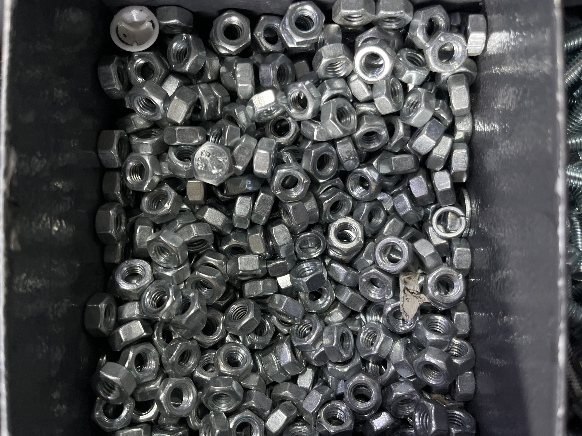 The image shows a close-up, overhead view of a bin filled with hexagonal nuts. The nuts are made of metal and appear to be silver or grey in color. They are of various sizes and are piled loosely in the bin. Some of the nuts are partially obscured by others. In the upper right corner of the image, there is a small white plastic object with a smiling face design. The background of the image is dark and appears to be the inside of the bin or a shelf. The overall impression is of a large quantity of hardware.