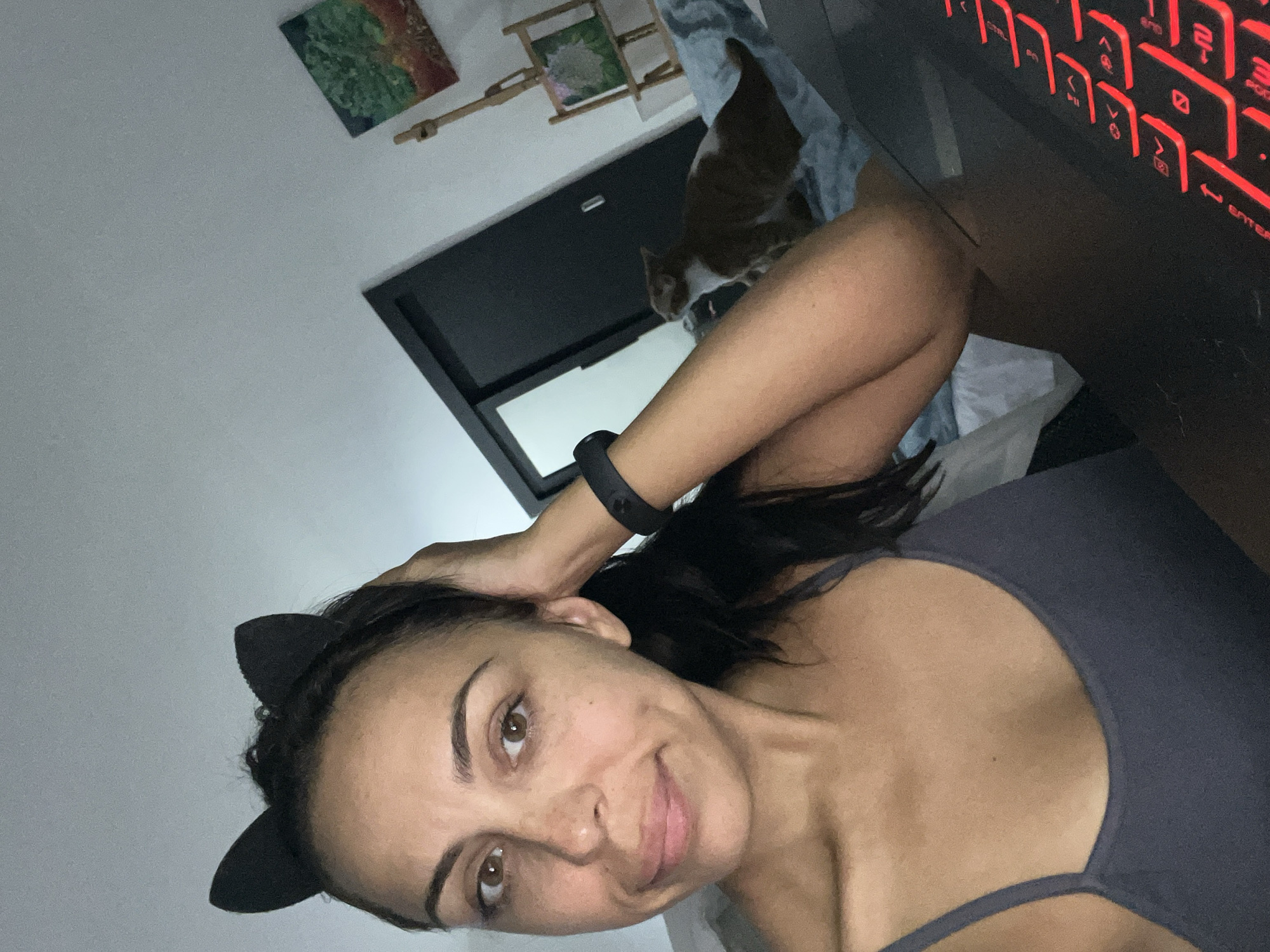 A selfie captured in an indoor setting in Kuta Selatan, Indonesia, features a woman and a cat. The woman, with light skin and dark hair pulled back, looks directly at the camera with a calm, slightly smiling expression. She is wearing a dark grey tank top and a black cat ear headband. A black fitness tracker is visible on her left wrist, and her hand is near her head, possibly adjusting her hair or the headband.
In the background, an orange and white cat is seen lounging on what appears to be a bed or furniture. The room has light-colored walls, adorned with a small painting featuring green and reddish-brown abstract patterns, and a wooden decorative item. A dark-framed door or window is also visible behind the cat. To the right, a black keyboard with bright red backlighting is partially visible, showing keys like "ESC", "TAB", "CAPS", "SHIFT", "ENTER", and numerical keys (1-0). The overall low light in the room, highlighted by the keyboard's glow, suggests the photo was taken during the evening or at night.