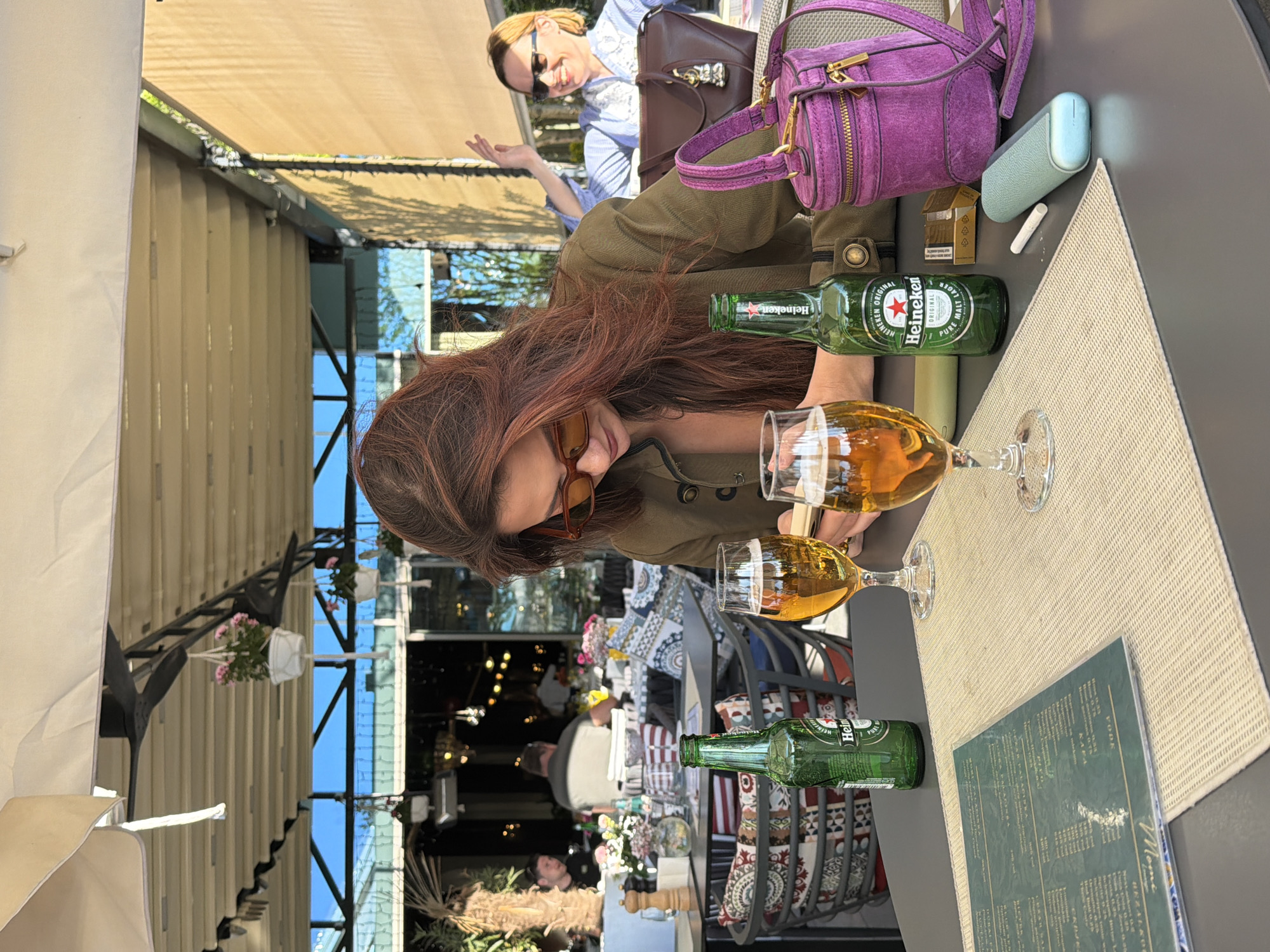 The image captures an outdoor cafe setting in Belgrade, Serbia. A young woman with auburn hair, wearing a green jacket and sunglasses, is seated at a table. On the table, there are two half-full beer glasses, two bottles of Heineken beer, a light blue vape device, an open pack of cigarettes, and a menu. A small purple purse is also on the table.
In the background, another woman is seated at a nearby table, wearing sunglasses and a light blue top. The cafe is partially covered by an awning with hanging flower baskets. Inside the cafe, blurred figures suggest other patrons. The setting implies a leisurely social gathering at a restaurant or bar in Belgrade.