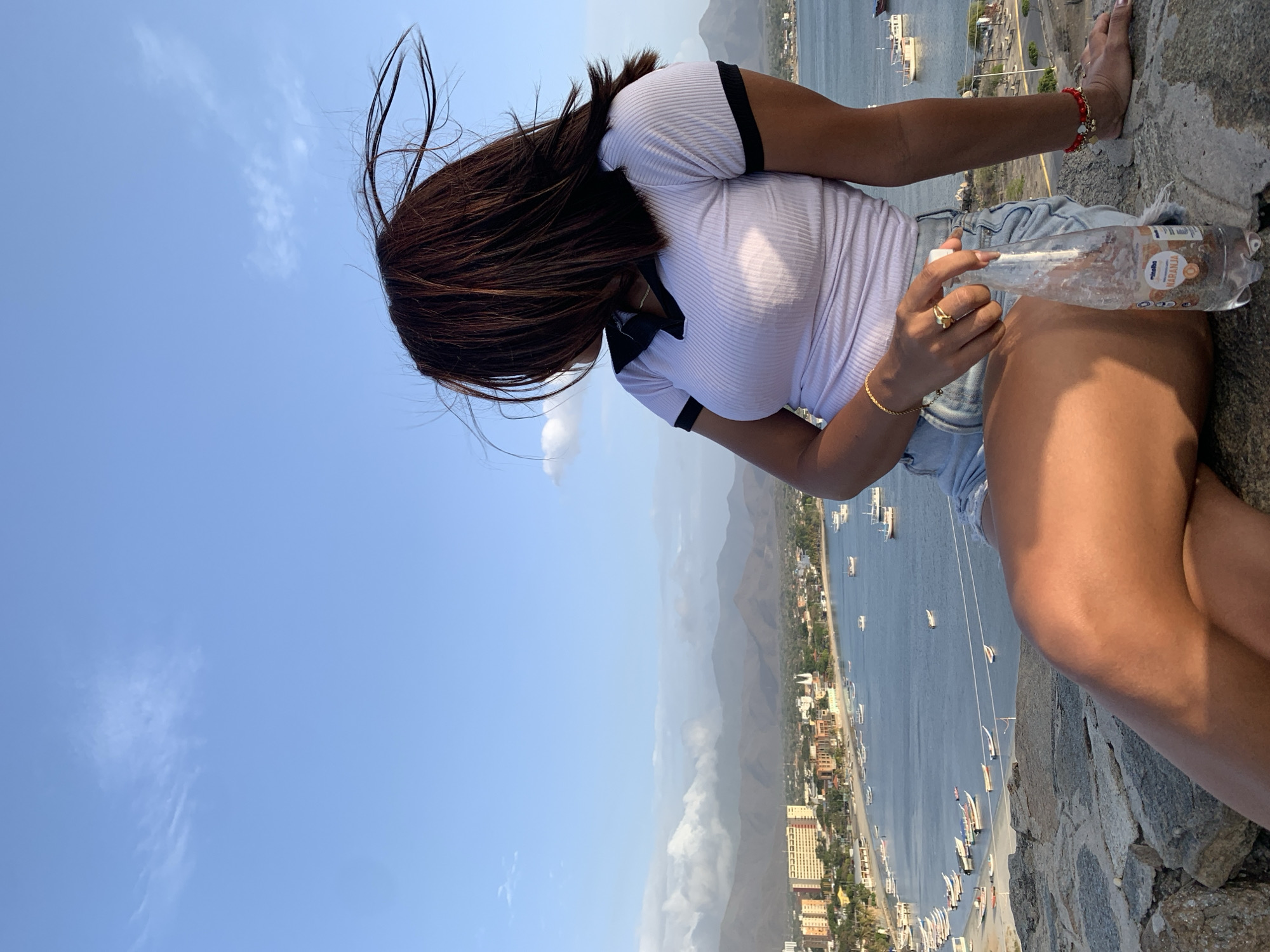 A high-angle, side-view image captures a woman sitting on a rocky ledge, overlooking a coastal landscape. The woman, with long, dark, windswept hair, is seen from her left side and back. She wears a white ribbed t-shirt with black trim on the collar and sleeves, and light blue denim shorts. Her left hand displays a gold ring and a gold chain bracelet, along with a red beaded bracelet. In her right hand, she holds a clear plastic water bottle with some ice and condensation. Visible text on the bottle includes "Albalba" and "NARANJA".
The foreground consists of textured grey and brown rocks where she is seated. Below her, a wide panoramic view unfolds, revealing a bustling coastal town situated along a calm body of water, likely a bay or ocean. Numerous boats, yachts, and smaller vessels are dotted across the water. Buildings of varying sizes line the shoreline, extending towards distant mountains that dominate the horizon, partially shrouded in atmospheric haze. The sky above is a clear, vibrant blue with a few scattered, wispy white clouds. The bright illumination suggests it is daytime, likely late morning or afternoon. The scene depicts a serene moment of observation or rest, with the windswept hair indicating a breezy environment. The overall setting is consistent with a scenic outlook in a coastal region like Juan Griego, Venezuela.