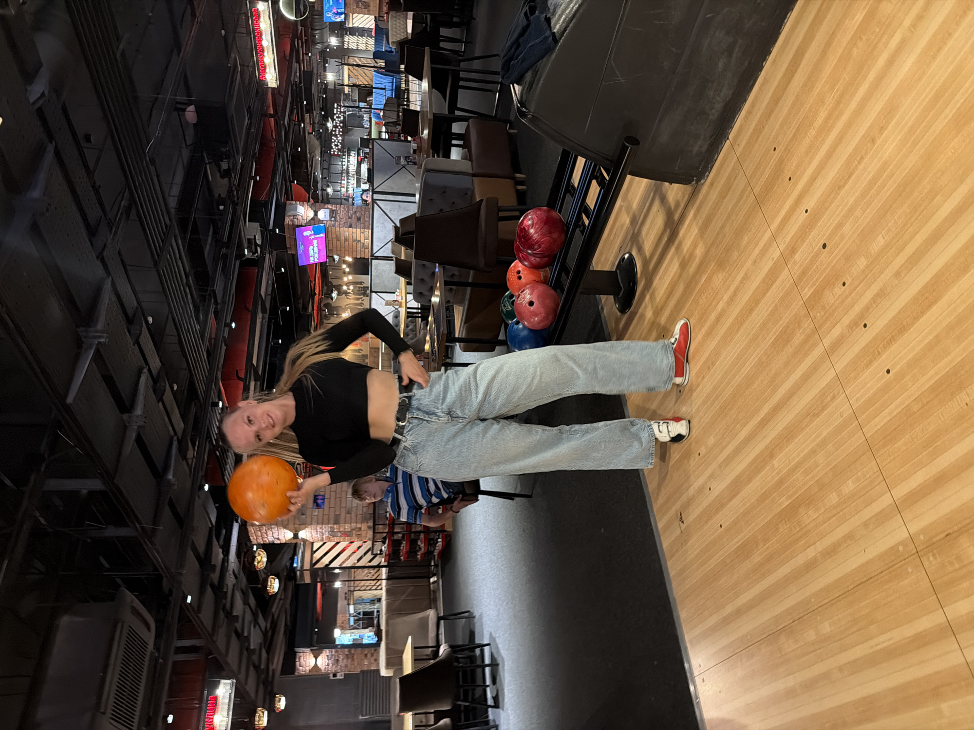 An adult female, appearing to be in her late teens or early twenties, stands on a wooden bowling lane, holding an orange bowling ball. She is wearing a black long-sleeved crop top, light blue wide-leg jeans, a black belt, and multi-colored sneakers. She is looking towards the camera and smiling. To her right, several bowling balls are arranged on a rack, including red, blue, and green ones. The background shows a bowling alley with tables, chairs, brick walls, and various signs and lights, consistent with an entertainment venue. The image was taken in the city of Izhevsk, Russia.
