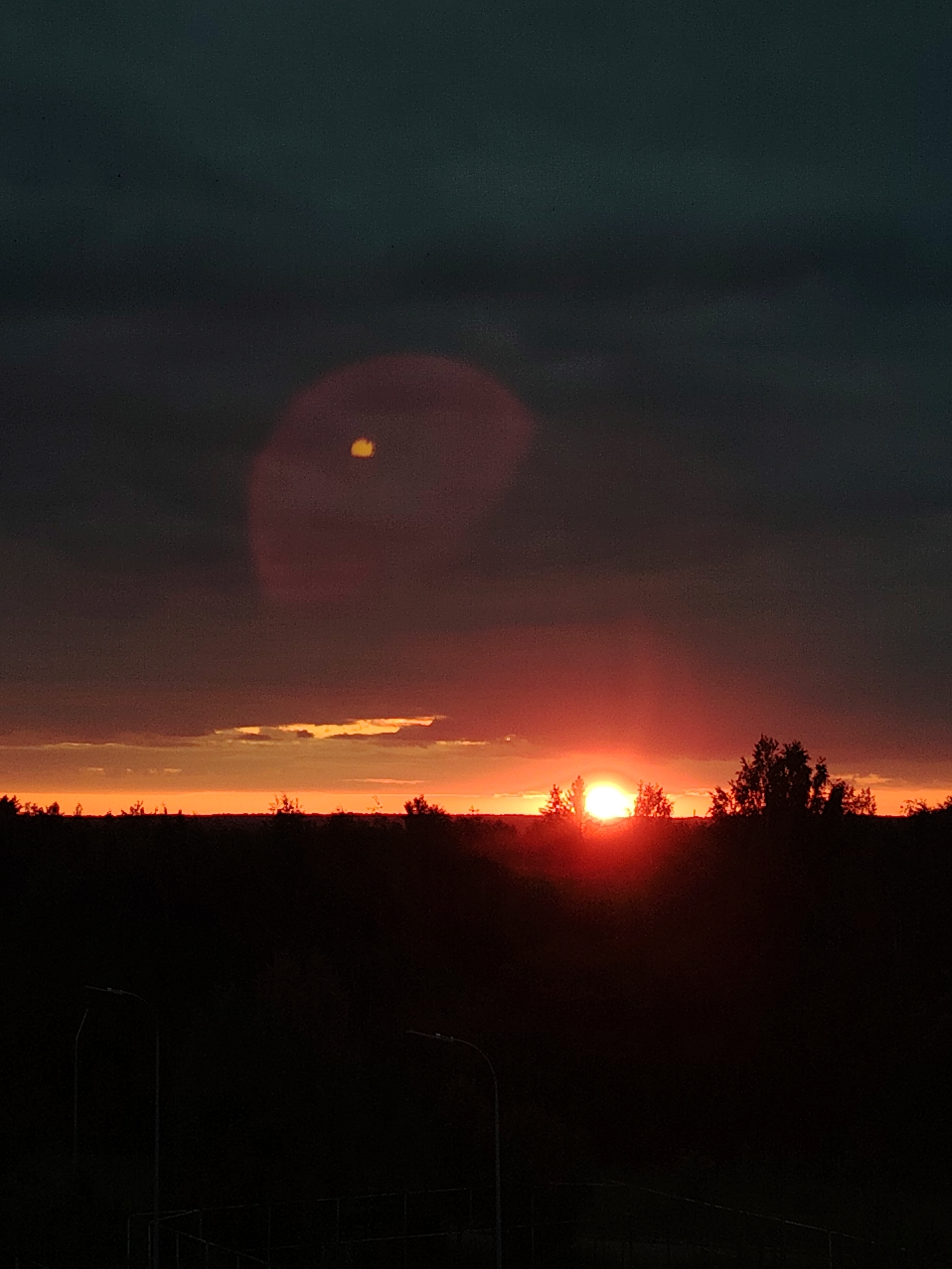A silhouette of trees and a dark landscape is visible against a dramatic sunset sky. The sun, a bright orange sphere, is partially obscured by a silhouetted tree line. The sky above transitions from a vibrant orange and red near the horizon to a deep, dark teal at the top. A faint, circular lens flare, colored red with a yellow-orange spot in the center, is present in the upper portion of the sky. In the foreground, two dark silhouettes of streetlights are visible on the left and right sides of the frame, positioned over a dark, unidentifiable structure. The scene appears to be an outdoor landscape, possibly on the outskirts of a city.