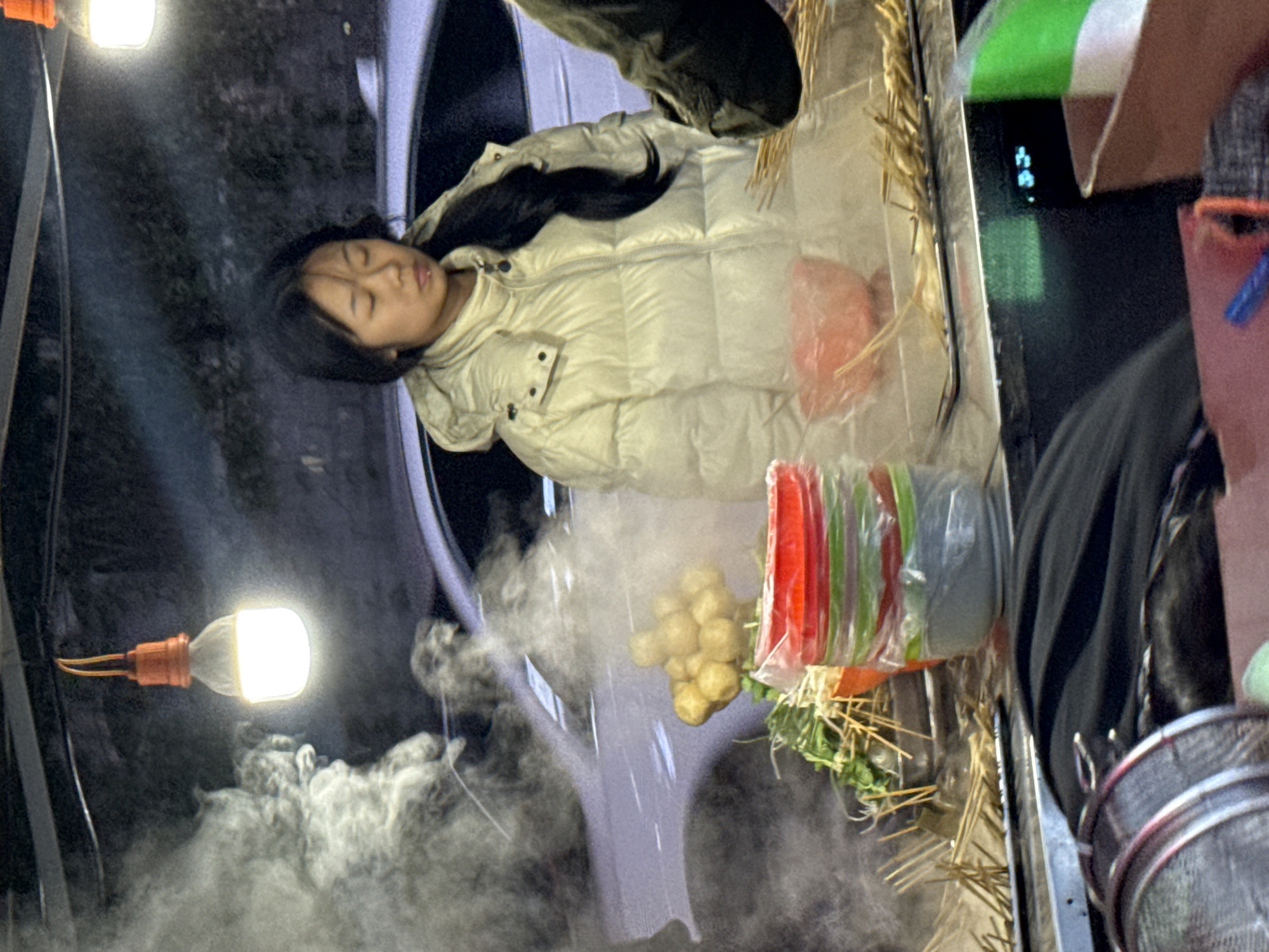 The scene shows a woman in a white puffer jacket resting her head inside a car parked near a street food vendor's setup. The vendor's stall includes a variety of food items on skewers, covered in plastic wrap. Steam or smoke is visible near the stall. A light bulb is positioned near the smoke. The location is Zhongtun, China.
