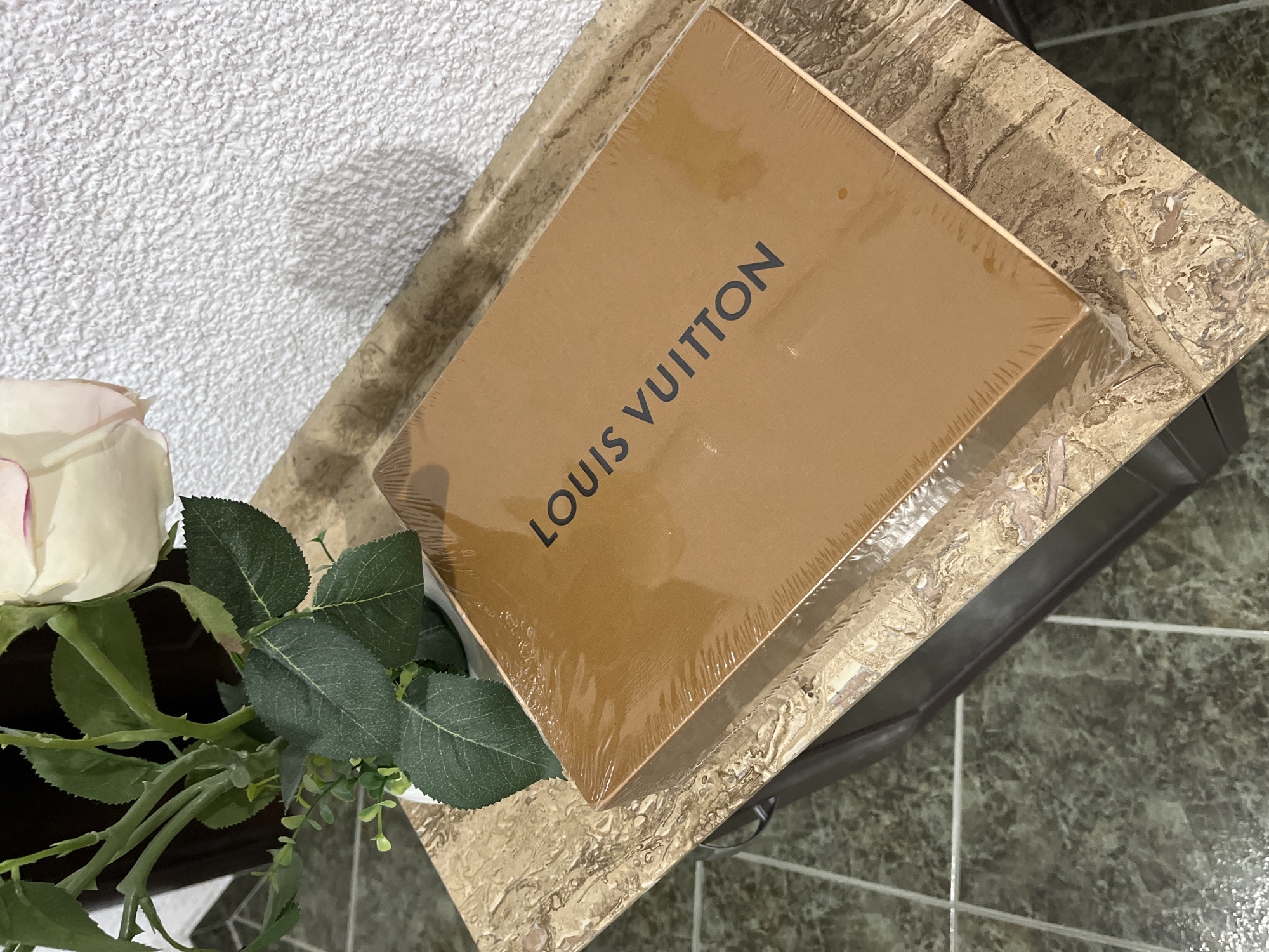 The image shows a brown Louis Vuitton box on a marble-patterned surface next to a vase of white roses and green foliage. The background includes a textured white wall and green tiled flooring. The image does not provide information about the location in the city of Zlatograd, Bulgaria.