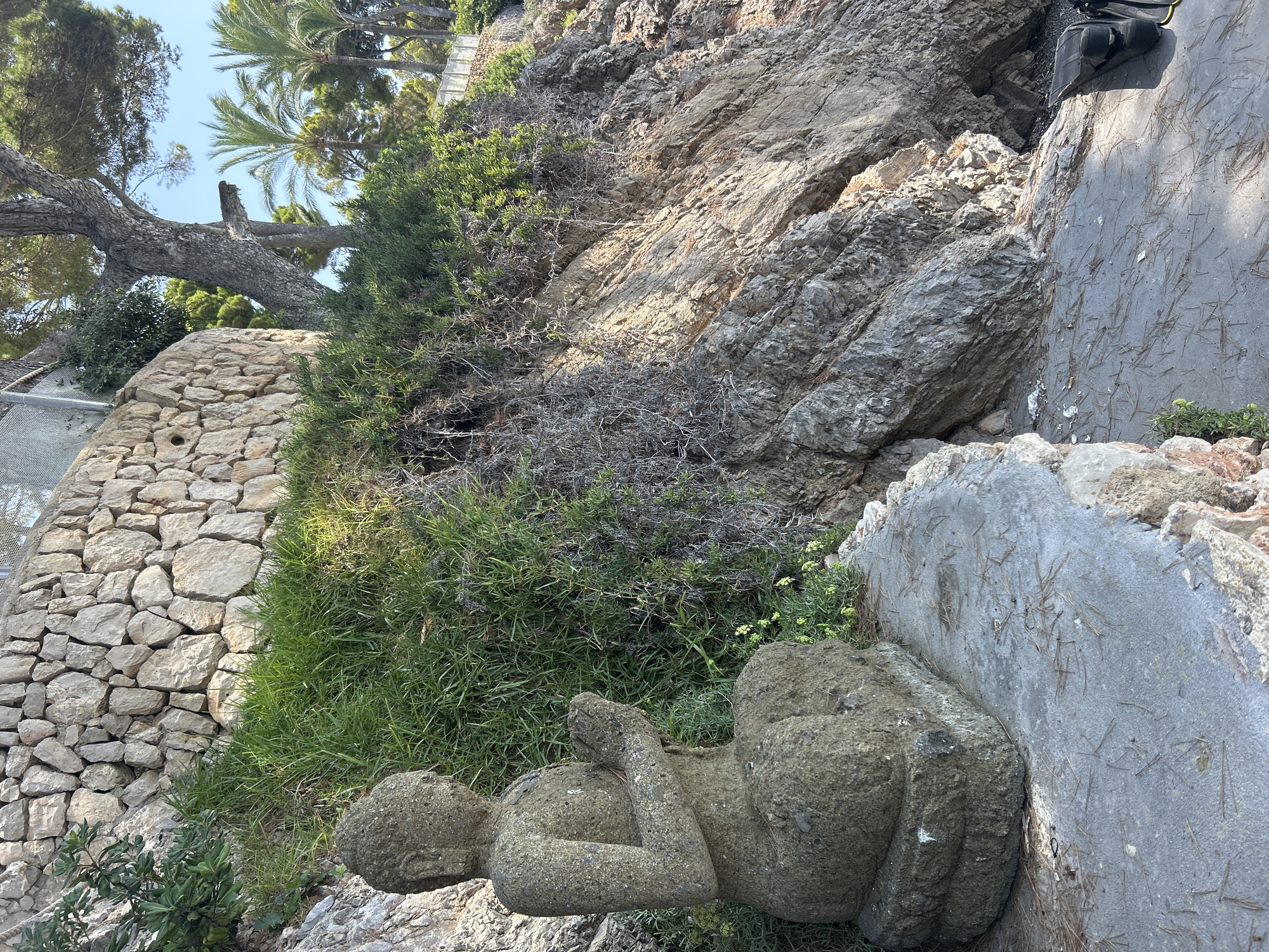 The image depicts a weathered stone statue of a meditating Buddha in a seated position, resting on a flat stone platform. The statue is positioned outdoors, with a rocky cliff face and lush green vegetation in the background. A stone wall is visible on the upper left side of the image, and a large tree with pine needles scattered around its base is present on the right. Palm trees and a clear blue sky suggest a warm, sunny climate. The overall scene evokes a sense of tranquility and natural beauty. The location is identified as Costa Des Pins, Spain.