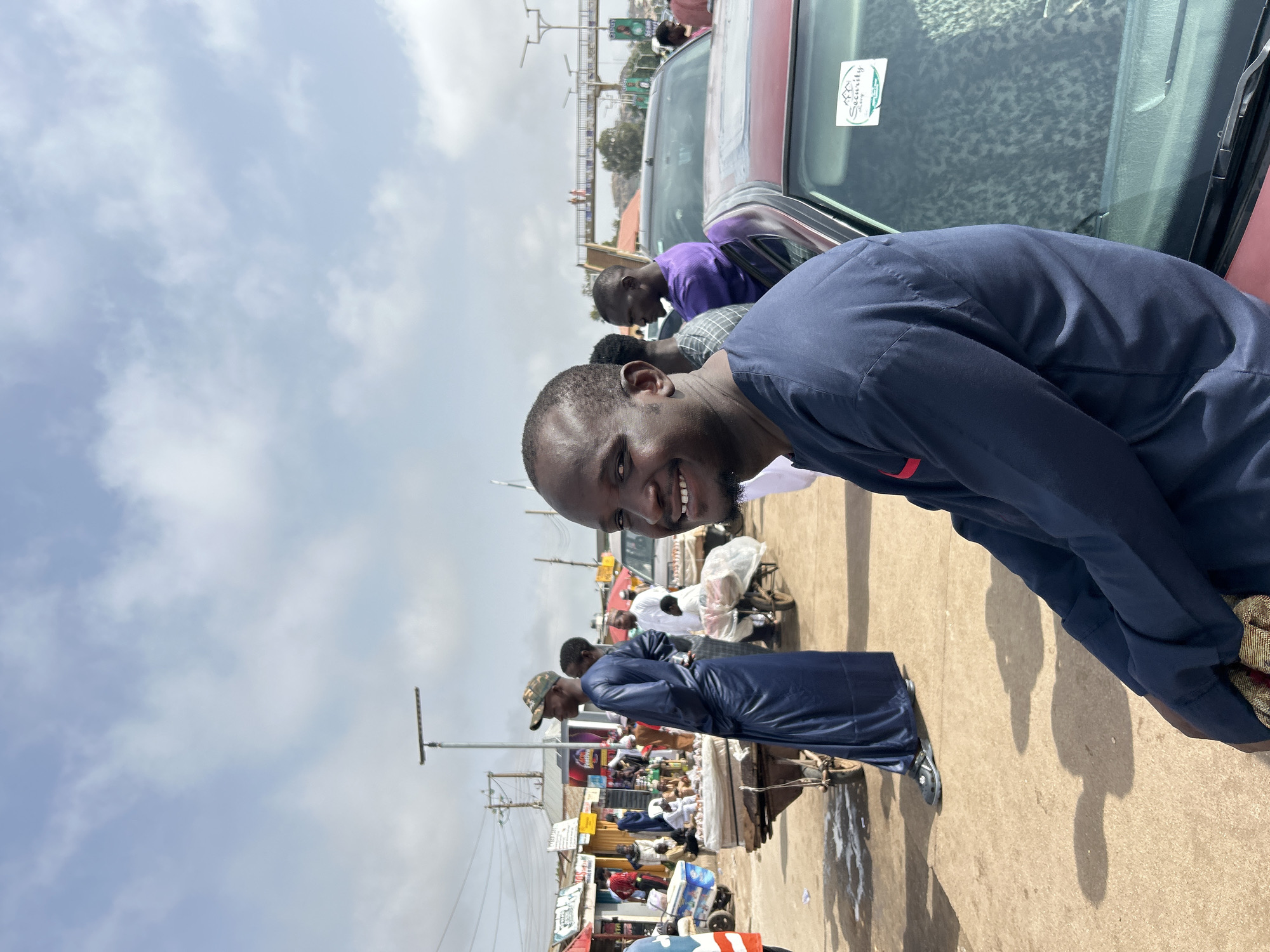 The image is taken in a busy area that appears to be a market in Jos, Nigeria. In the foreground, a man smiles at the camera while leaning towards a maroon-colored vehicle. A sticker is visible on the car window. Behind him, other people are visible in the background, some wearing casual clothes. Stalls and displays of goods can be seen lining the area. Power lines and light poles are visible overhead against a backdrop of a cloudy sky.