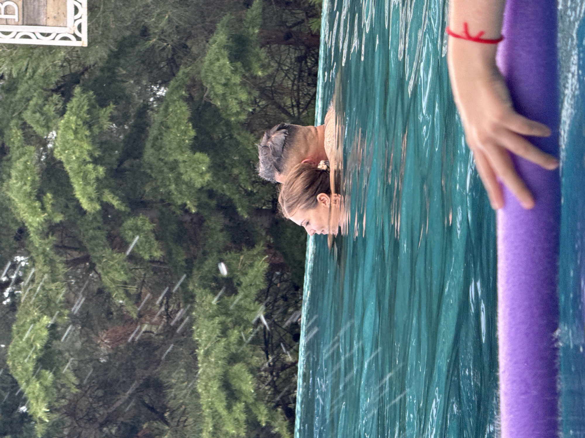 In the city of Yalta, Ukraine, two individuals, a man and a woman, are submerged in a turquoise swimming pool. The woman is positioned on the left, facing left, with her head and shoulders visible above the water. The man is to her right, with his head and shoulders also above the water. They appear to be embracing or in close proximity. The pool is an infinity pool, with the water extending to the edge and a backdrop of lush green pine trees. The sky is overcast, with visible rain or water droplets in the air. In the foreground, a person's hand is visible resting on a purple pool noodle. A decorative white sign with the letter 'B' is visible in the upper right corner, partially obscured by trees.