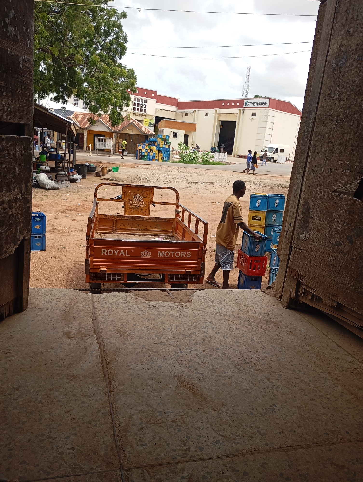 A young Black male, wearing a yellow t-shirt with a large number "1" printed on the back and denim shorts, is standing to the right of a small orange three-wheeled motorized cart, stacking blue plastic crates. He is holding a red plastic crate in his hands, placing it on top of a stack of blue crates. The cart, labeled "ROYAL MOTORS," is empty except for some items in the bed. Behind the man and cart are stacks of blue plastic crates, some labeled "STAR." A sandy, dirt ground covers the scene. In the background, several buildings are visible, including one with the text "KATYEN HOUSE" prominently displayed. A white van is parked near the building. To the left, a covered market stall with various pots and containers is present. Several people are walking in the distance. The sky is overcast with clouds. The overall setting appears to be an outdoor market or commercial area in Shendam, Nigeria.