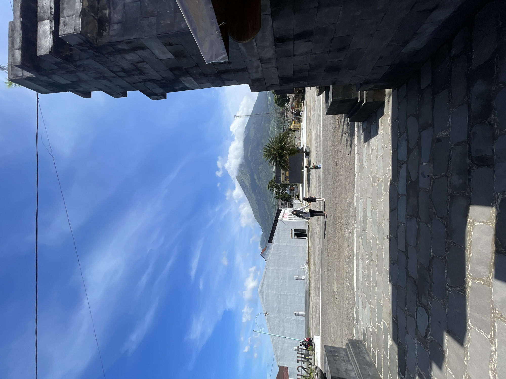 The image shows an outdoor scene in Kledung, Indonesia, with a view framed by the dark stone architecture of a building. In the distance, a mountain is visible, partially covered by clouds, set against a bright blue sky with wispy clouds. A building of light grey color, and with a simple design, is seen in the center, while two people are walking away from the camera across a paved area. A palm tree is also visible.