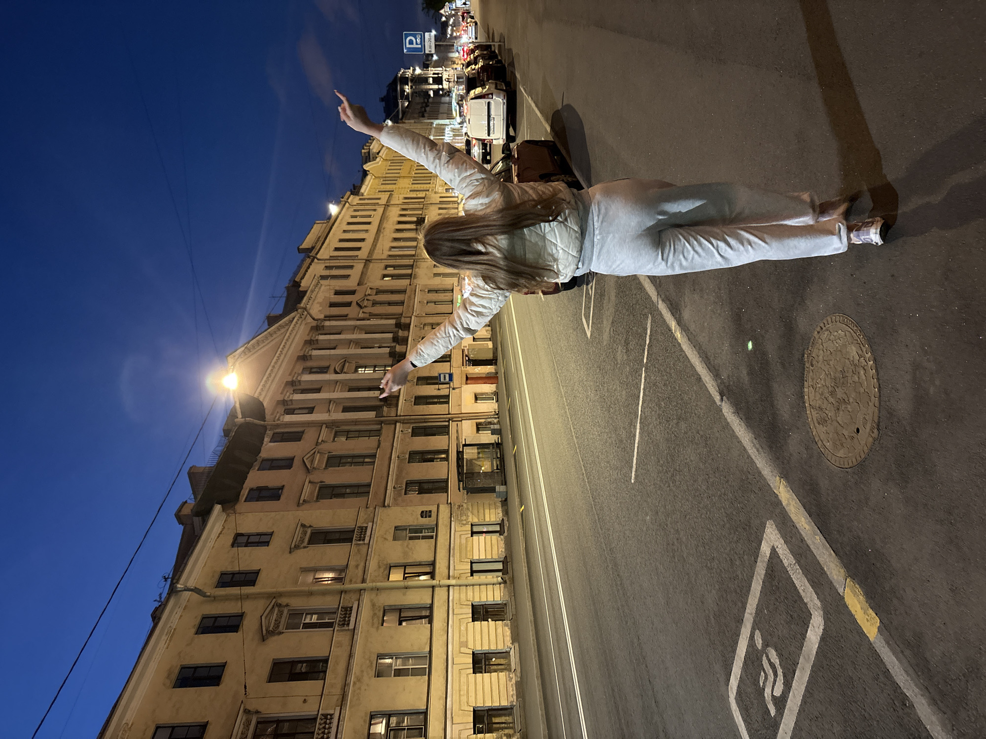 A woman is lying on her back on the street. She has long blonde hair and wears a white jacket and pants. She has both arms outstretched and her index fingers pointed upwards. The background shows a typical Saint Petersburg, Russia street scene at night. An old yellow building with many windows is visible on the left, and there is a dark blue sky above. Cars, traffic lights, and street signs are visible in the distance on the right. There is a manhole cover in the pavement near the bottom of the frame.