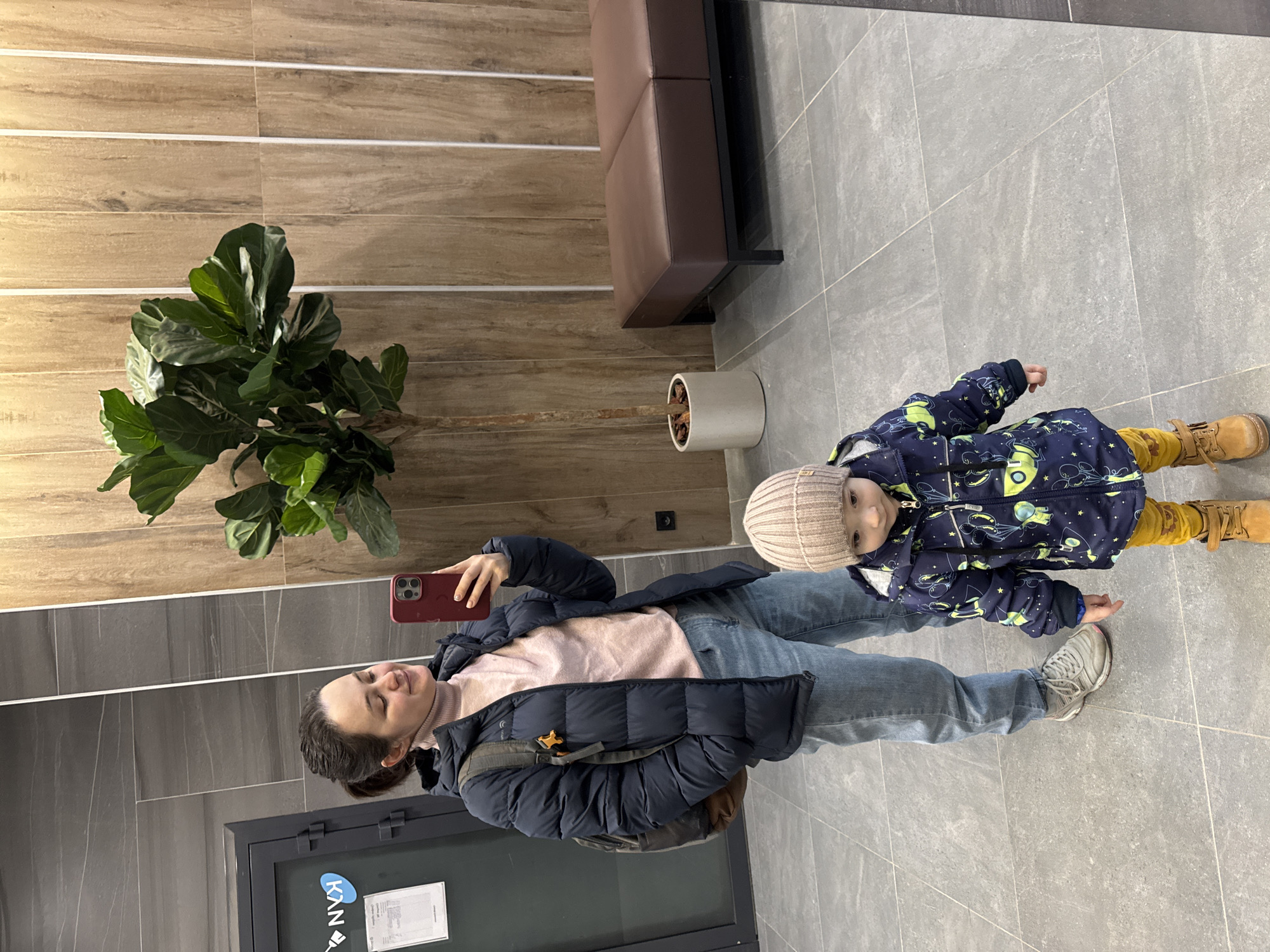 A woman and a child are indoors. The woman is holding a red phone and appears to be taking a selfie. She is wearing a dark blue puffer jacket, a light pink turtleneck sweater, and light blue jeans. The child is wearing a dark blue printed jacket, yellow pants, a light-colored knit hat, and tan boots. A large plant is visible on the left side of the image. A bench is situated in the background. The floor is tiled. A door is partially visible to the left. According to the door sign, the location is in Izhevsk, Russia.