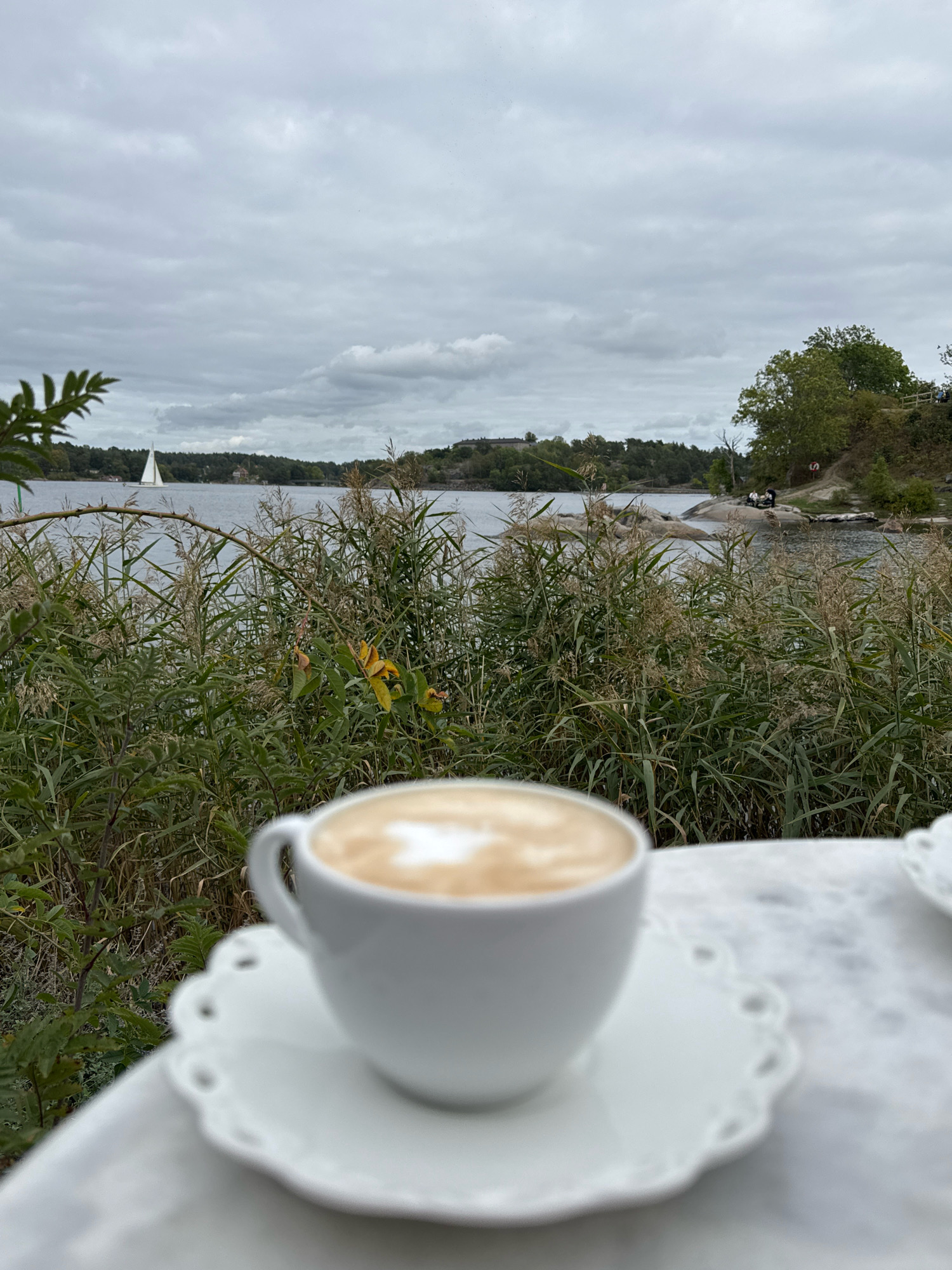 In Vaxholm, Sweden, a white cup of coffee with latte art sits on a white saucer with a scalloped edge. The cup and saucer are placed on a white surface, possibly a table with a textured or patterned cloth. In the background, a calm body of water is visible, with tall reeds and green foliage in the foreground. A white sailboat is sailing on the water in the distance. The sky is overcast with clouds. Further in the background, a treelined shore with buildings and a few people are discernible on a rocky outcrop.