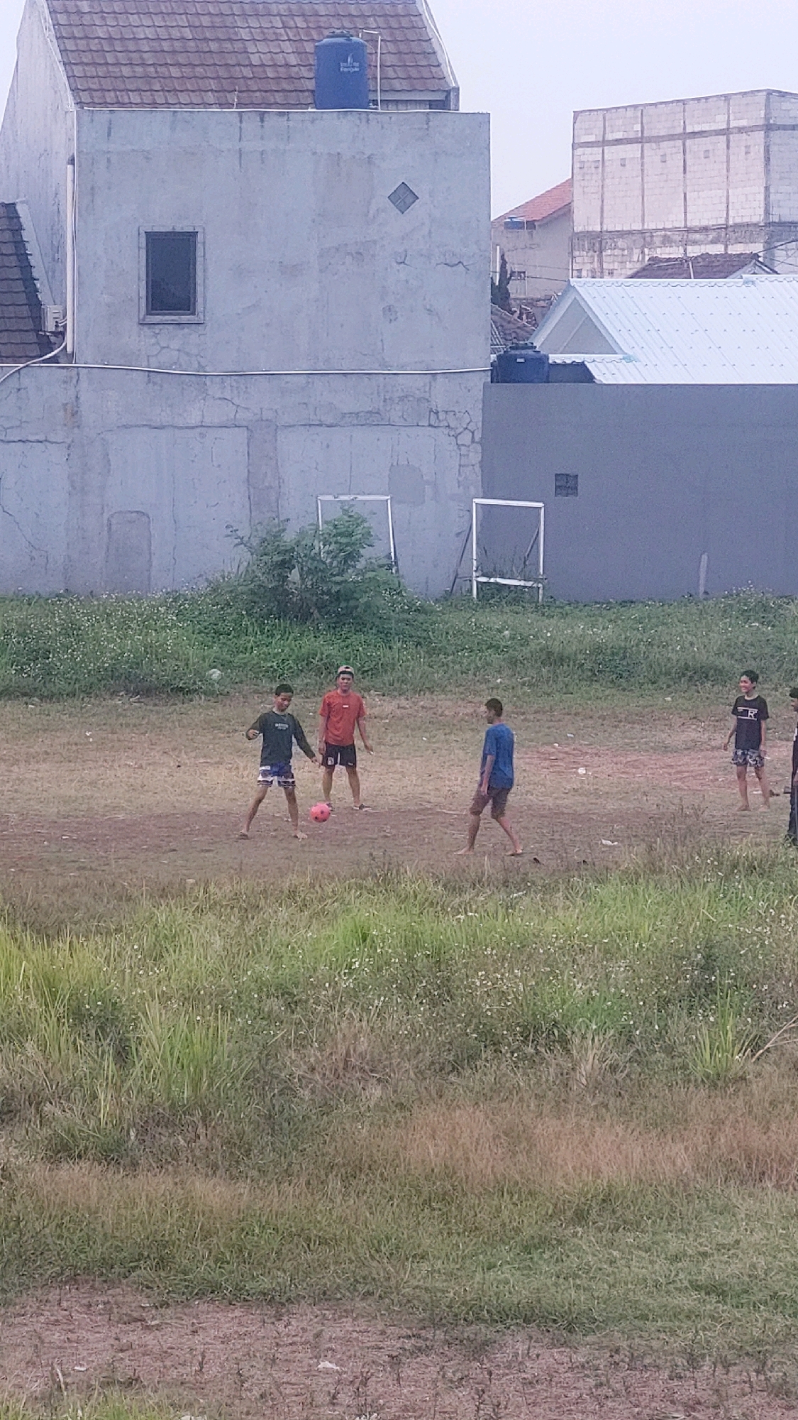 The image depicts a group of young boys playing soccer on a dirt field in Bojong Gede, Indonesia. The field is surrounded by tall grass and some scattered white wildflowers. In the background, there are buildings. One prominent building is a light gray, multi-story structure with a brown tiled roof and a blue water tank on top. Another building in the background is a lighter color with a white metal roof and a dark blue water tank.
There are five boys visible in the image. One boy in a dark green shirt and dark shorts is dribbling a pink soccer ball. Another boy in a red t-shirt and black shorts is standing nearby, looking at the ball. A third boy in a blue t-shirt and brown shorts is also engaged in the game. Two other boys are visible on the right side of the frame; one in a black t-shirt with an "R" logo and patterned shorts, and another in a black shirt and shorts. They appear to be observing or participating in the soccer game. The overall scene suggests a casual outdoor activity taking place in a semi-urban or suburban environment.
