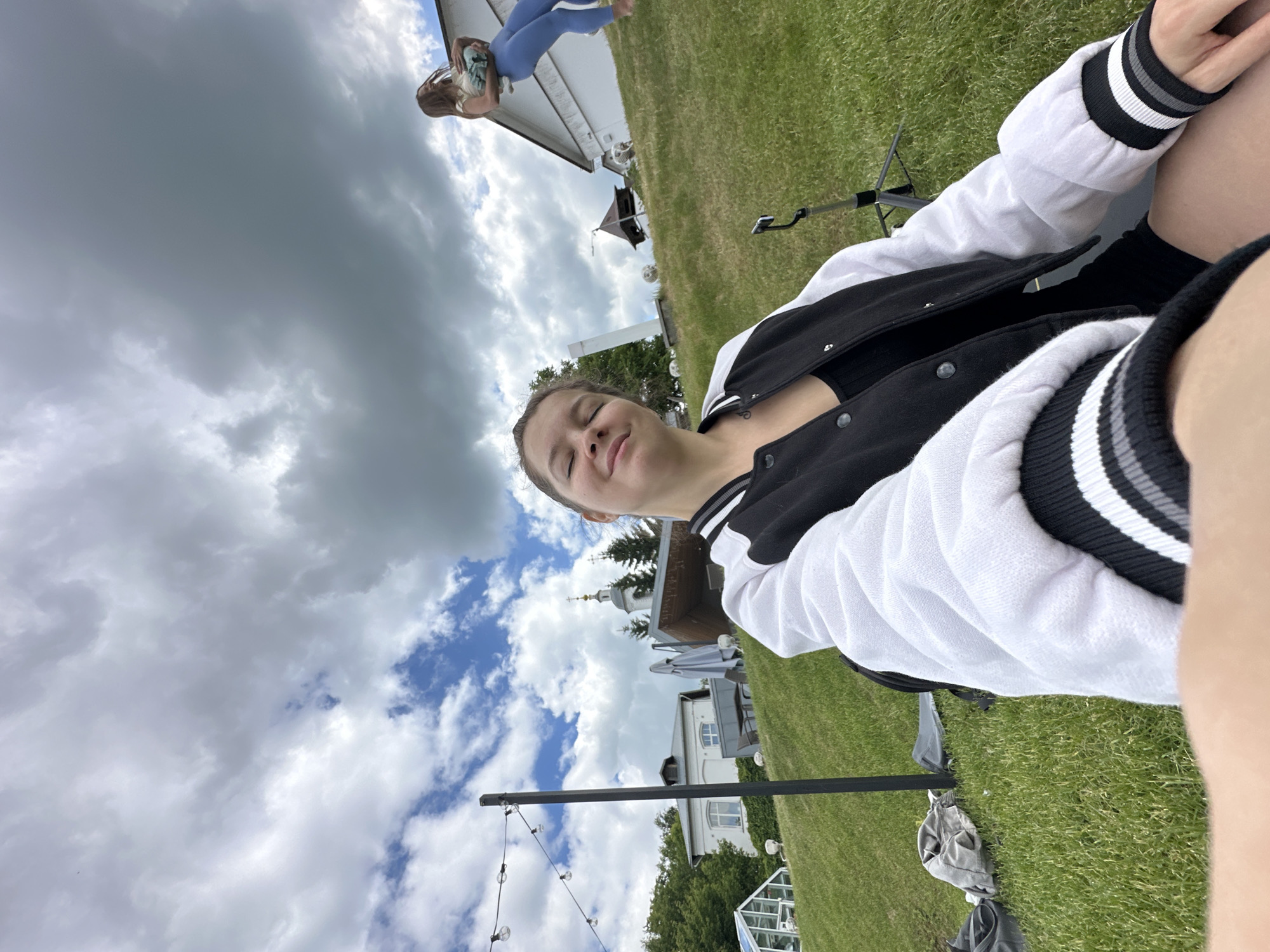 The image captures an outdoor scene on a partly cloudy day, likely in Vladimir, Russia. The primary subject is a young woman, positioned on the right side of the frame, appearing to be seated or reclining. She has brown hair, her eyes are closed, and she wears a gentle smile, conveying a sense of peace or contentment. She is dressed in a black and white varsity-style jacket with striped cuffs, over a dark top.
In the upper left background, another person wearing blue pants and a light top is visible, standing on a white building, possibly a balcony or porch. The setting features a lush green lawn, suggesting a garden or park area. Prominent in the mid-left background are several buildings: a white two-story structure, and further back and slightly to the right, a building with a distinctive onion-domed roof crowned with a cross, characteristic of Russian Orthodox architecture, which serves as a strong location cue for Vladimir. Other structures, including what looks like an outdoor seating area with umbrellas and a small greenhouse, are also visible.
A black adjustable stand or tripod is situated on the grass in the middle ground, and a string of decorative lights hangs from a pole in the lower left foreground. The sky overhead shows a mix of heavy grey clouds and patches of blue sky with white clouds, indicating variable weather conditions. The overall activity seems to be a relaxed outdoor gathering or day out. No visible text is present in the image.