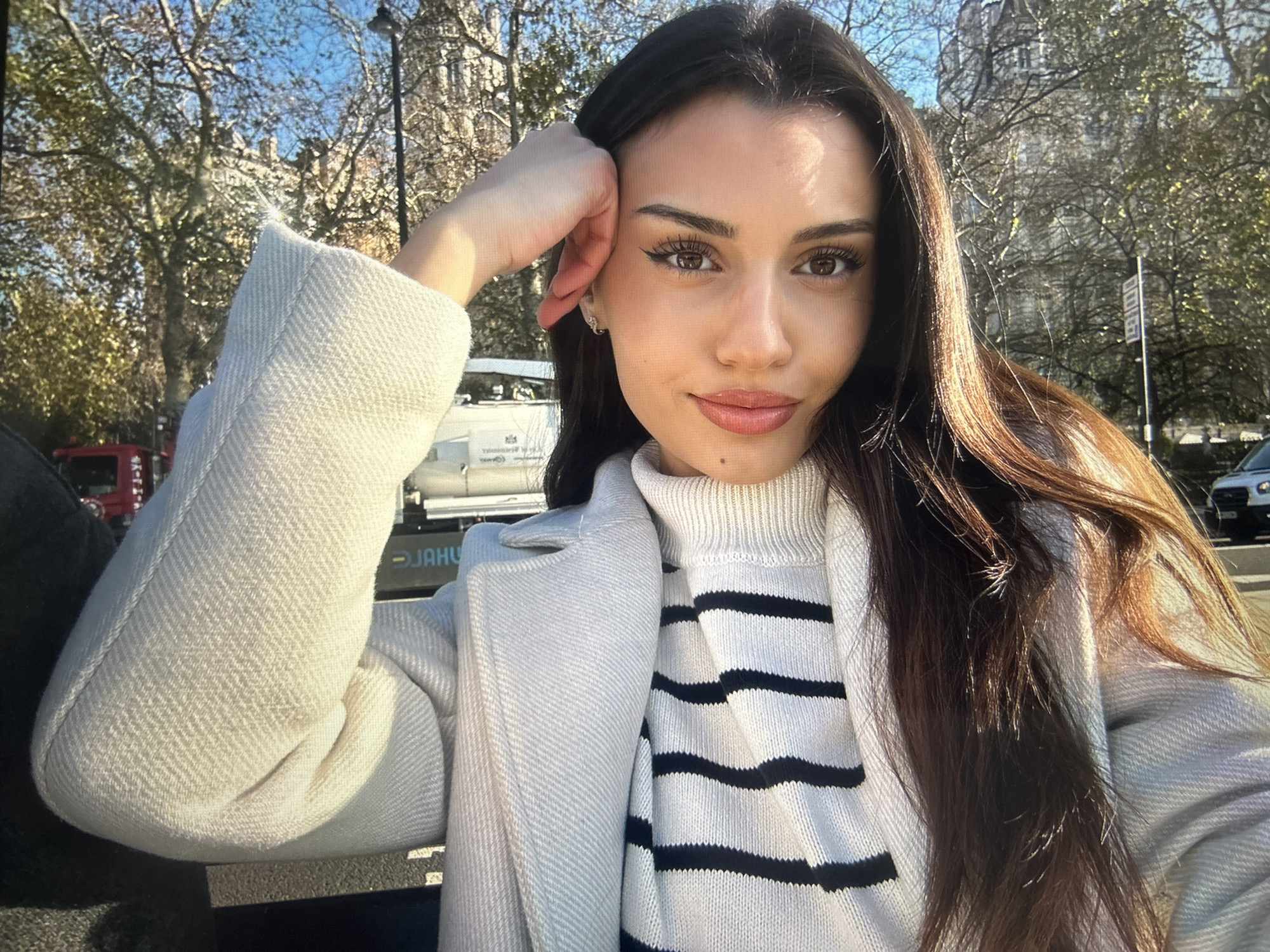 A woman with long brown hair is posing for a selfie outdoors, wearing a white turtleneck sweater with black stripes and a light-colored coat. She is resting her head on her hand. The background features trees, buildings, and a vehicle. The location is difficult to determine from the image alone, and cannot be identified as Gorna Oryahovitsa, Bulgaria.