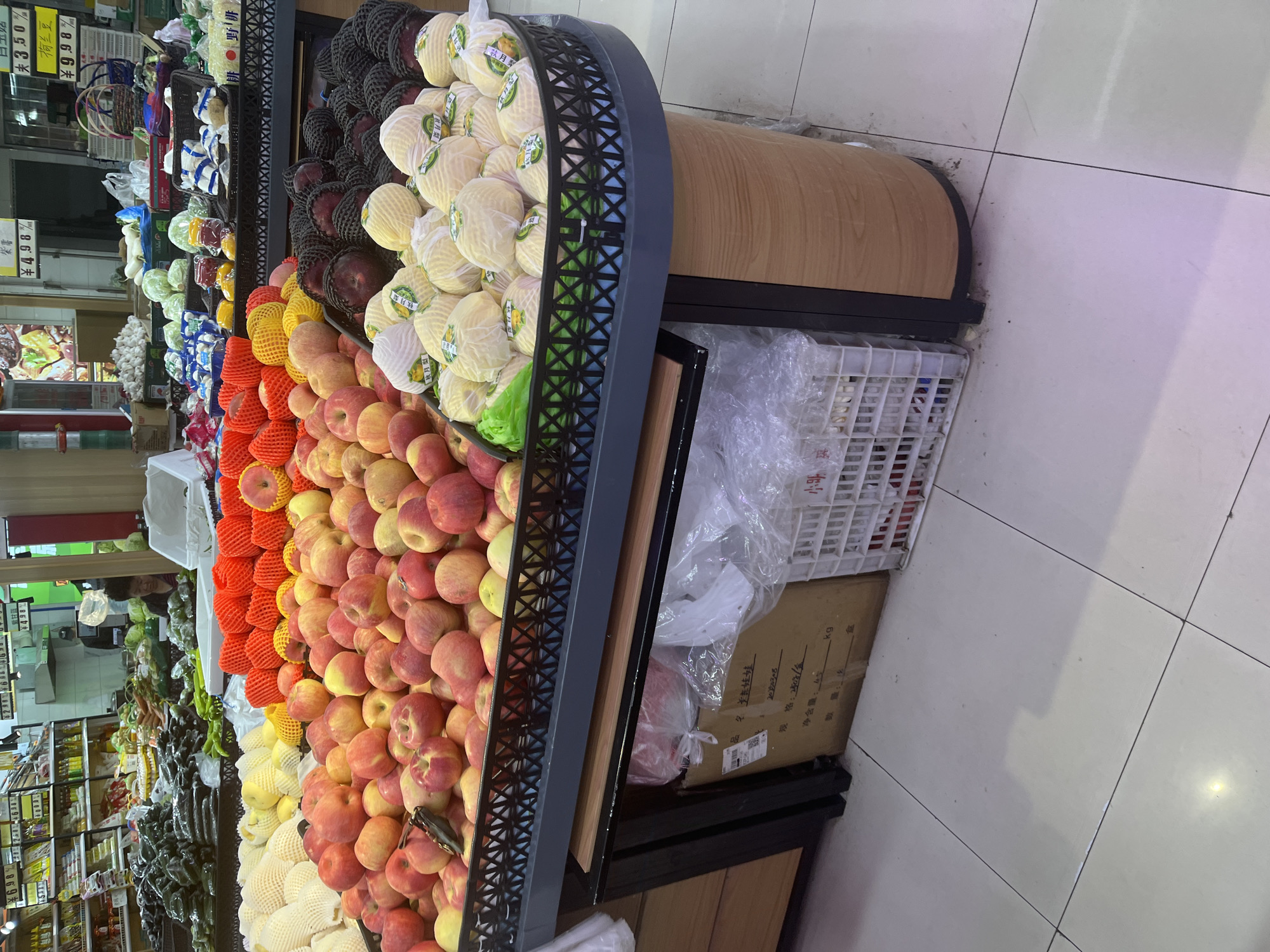 Here is a detailed description of the image:
**Scene:** A fruit display at a grocery store in Changchun City, China.
**Objects:**
* Various fruits are arranged in rows within a dark-colored, lattice-sided display stand. These fruits include:
* Apples, many packaged in red and yellow foam netting
* Pears (some packaged in black netting, some wrapped in white paper)
* Other fruits and vegetables visible further back.
* A brown wooden panel forms part of the display’s construction.
* A white plastic crate and a cardboard box are stored beneath the display stand.
* Shelves displaying various grocery items can be seen in the background.
* Price signs and labels are displayed.
**Environment:**
* The floor is tiled.
* The grocery store appears to be well-lit.
**Details from Text:**
* The text on the box mentions the specifications, stating a net weight of 4.5 kg, and a packaging date of 2020308.
* Price signs in the background show items like "Dutch beans," "Purple sweet potatoes."
